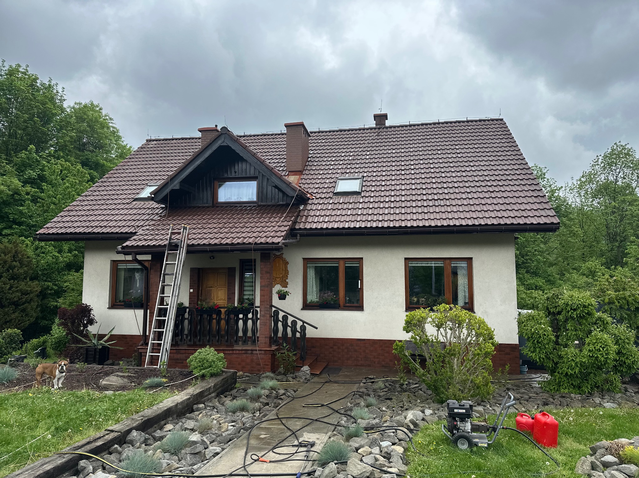 Dwupiętrowy dom z brązowym dachem i drabiną opartą o ścianę. Na trawniku myjka ciśnieniowa i dwa czerwone kanistry. Pochmurne niebo w tle.