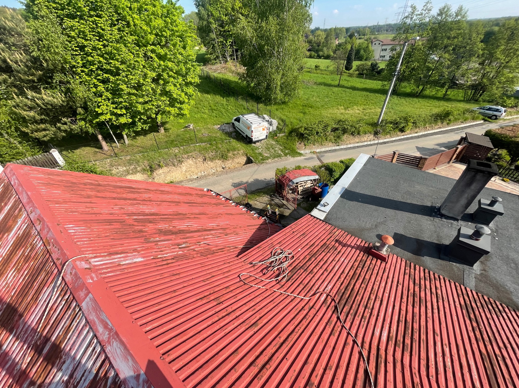 Czerwony, blaszany dach z widocznymi śladami korozji i częściowo odchodzącą farbą. Na dachu leżą liny i mała szczotka. W tle zieleń i budynki mieszkalne.