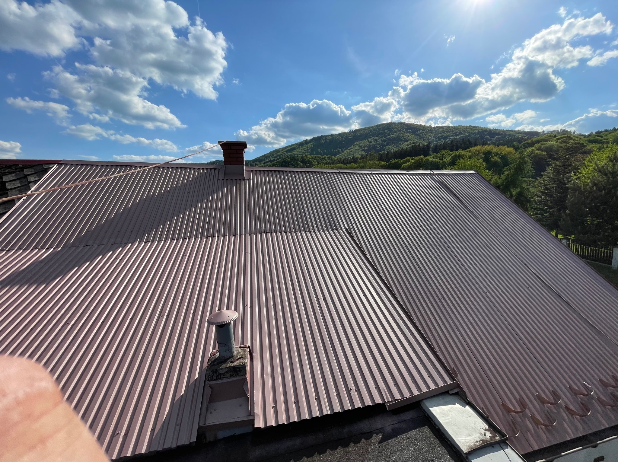 Falista blacha trapezowa na dachu w odcieniach brązu, z kominem i wentylacją, na tle błękitnego nieba z obłokami i zalesionego wzgórza.