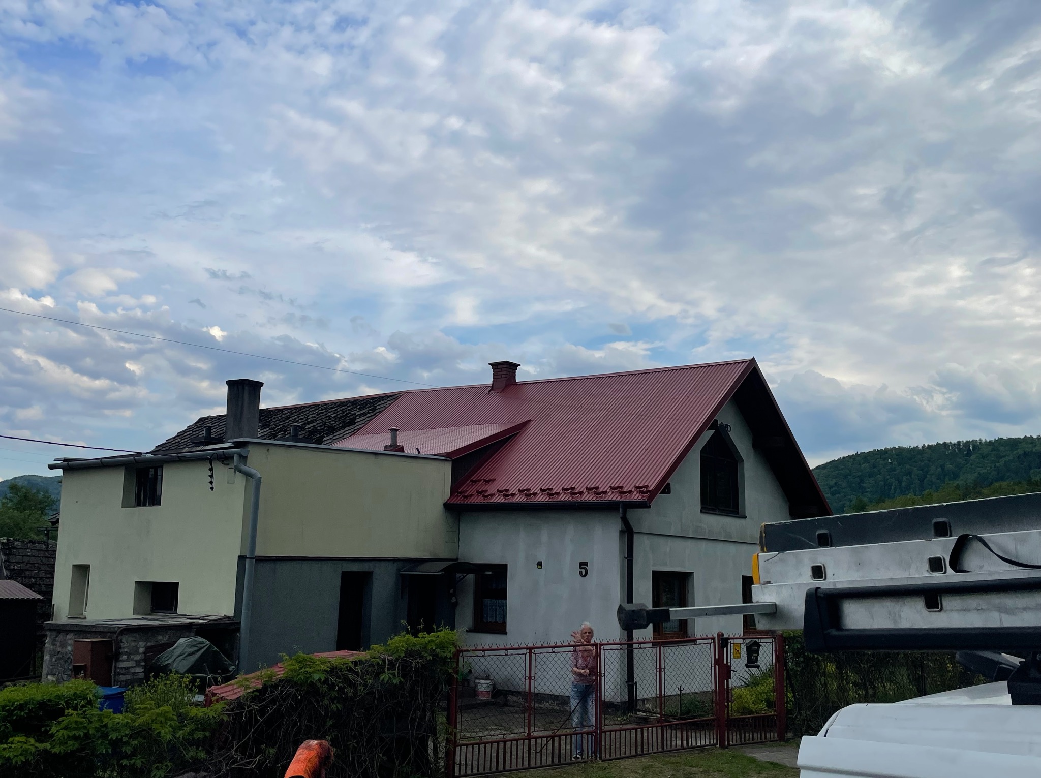 Dom z częściowo odnowionym dachem w kolorze czerwonym, widoczna drabina i osoba za ogrodzeniem. Połowa dachu przed renowacją. Pochmurne niebo w tle.