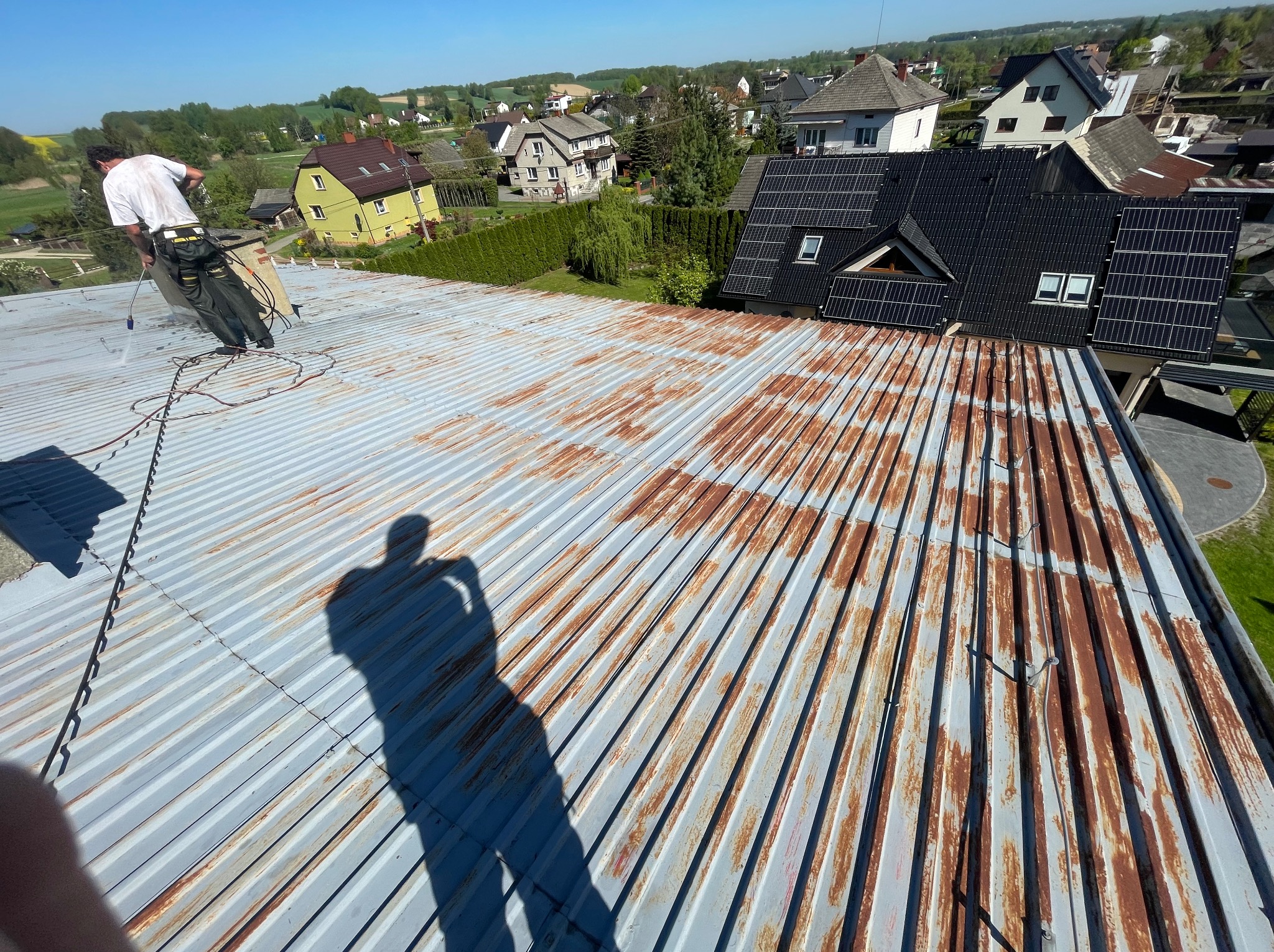 Pracownik w trakcie mycia zardzewiałego dachu z blachy falistej za pomocą myjki ciśnieniowej. Widoczny cień osoby robiącej zdjęcie, w tle zabudowa jednorodzinna.