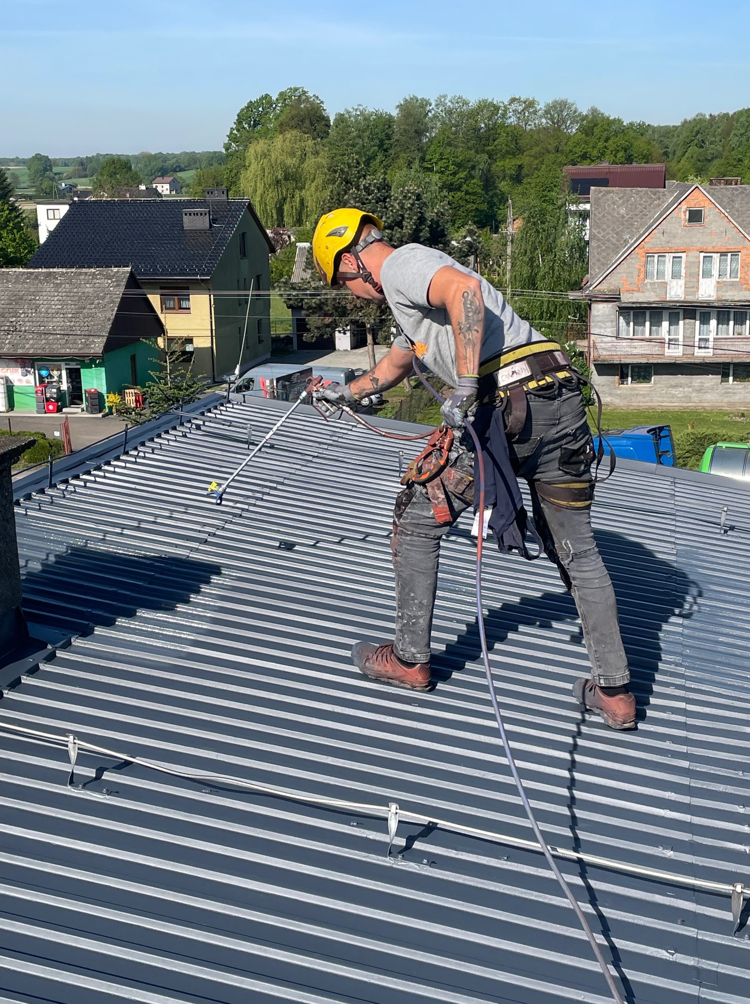 Pracownik w kasku, z zabezpieczeniami, maluje metalowy dach za pomocą pistoletu natryskowego. W tle zabudowa jednorodzinna i drzewa.