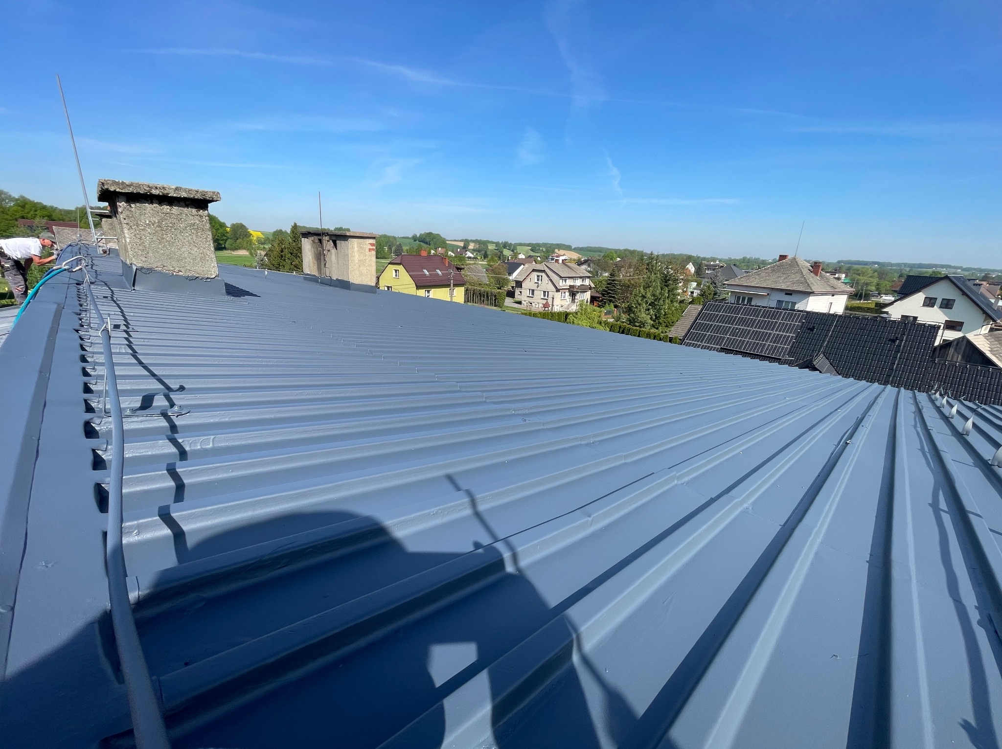 Pomarańczowo-szary dach z blachy trapezowej po malowaniu, widok z góry na tle zabudowy jednorodzinnej i błękitnego nieba. Prace dekarskie w toku, widoczny pracownik.