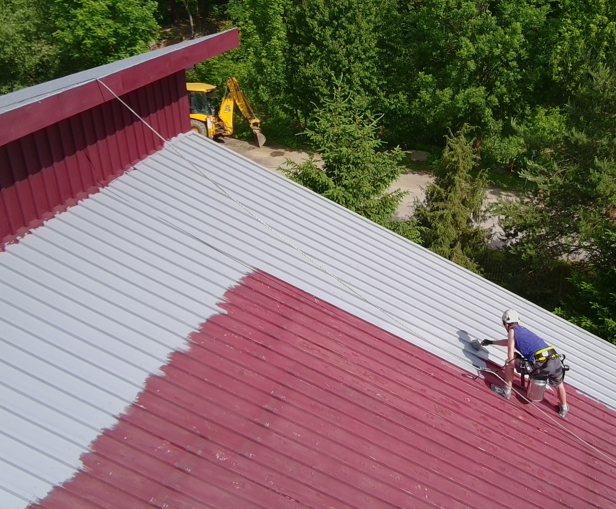 Częściowo pomalowany metalowy dach na czerwono i szaro. Pracownik z zabezpieczeniami maluje dach, w tle żółta koparka i zieleń drzew.