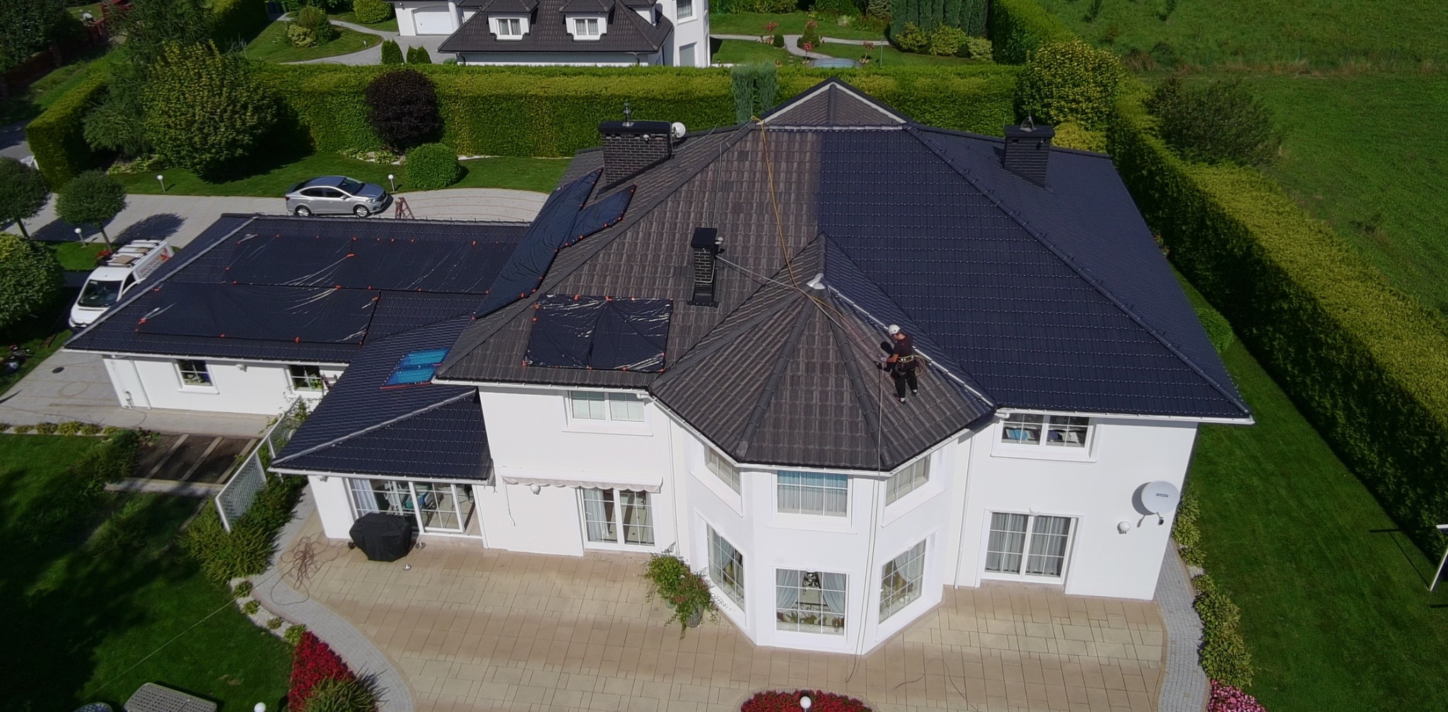 Częściowo pomalowany dach domu z białymi ścianami i pracownikiem w uprzęży, widok z drona, prezentujący postęp prac malarskich na tle zieleni.