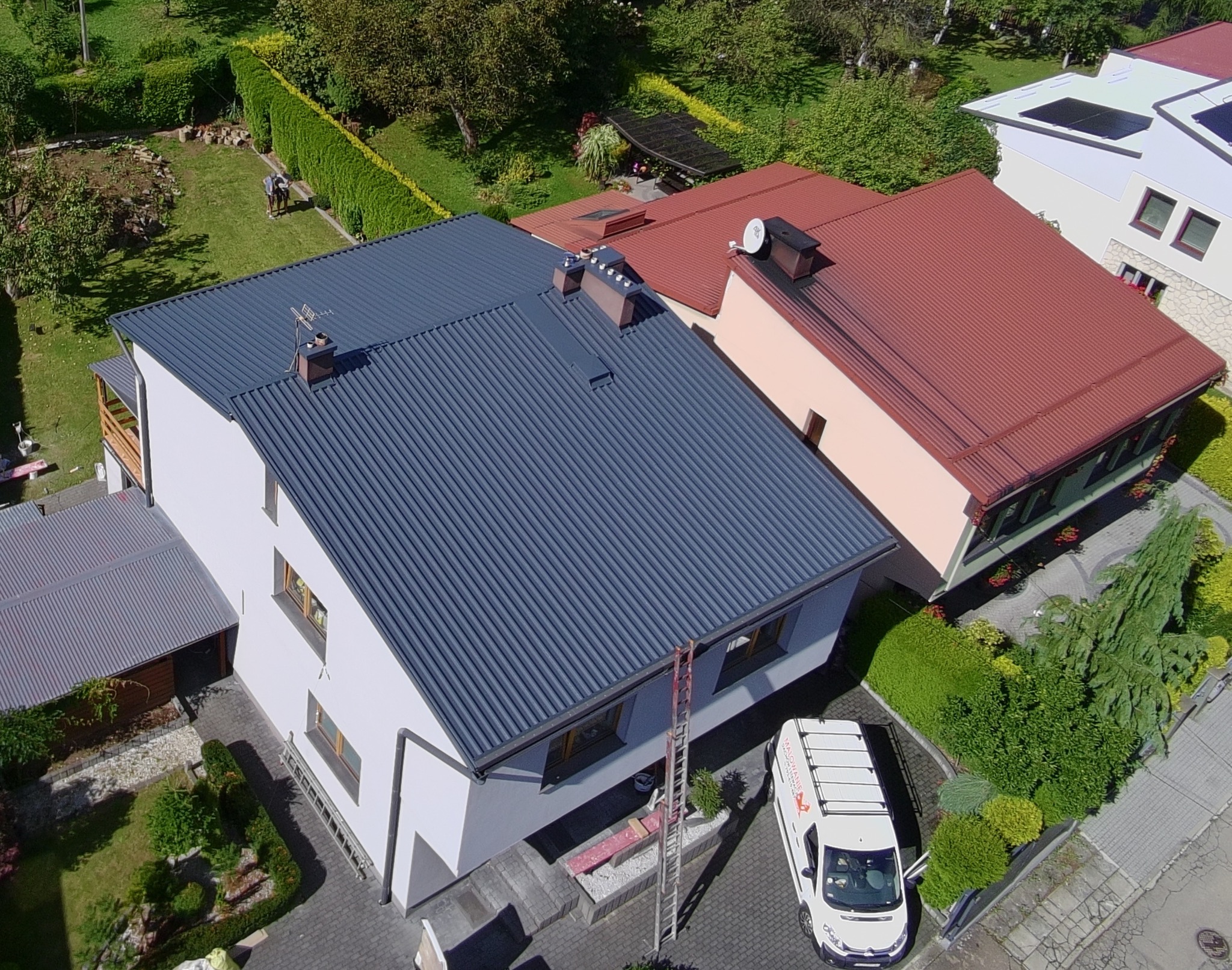 Widok z góry na dom z ciemnoszarym, odnowionym dachem z blachy trapezowej. Obok dom z dachem w kolorze ceglastym. Na podjeździe stoi bus firmy budowlanej.