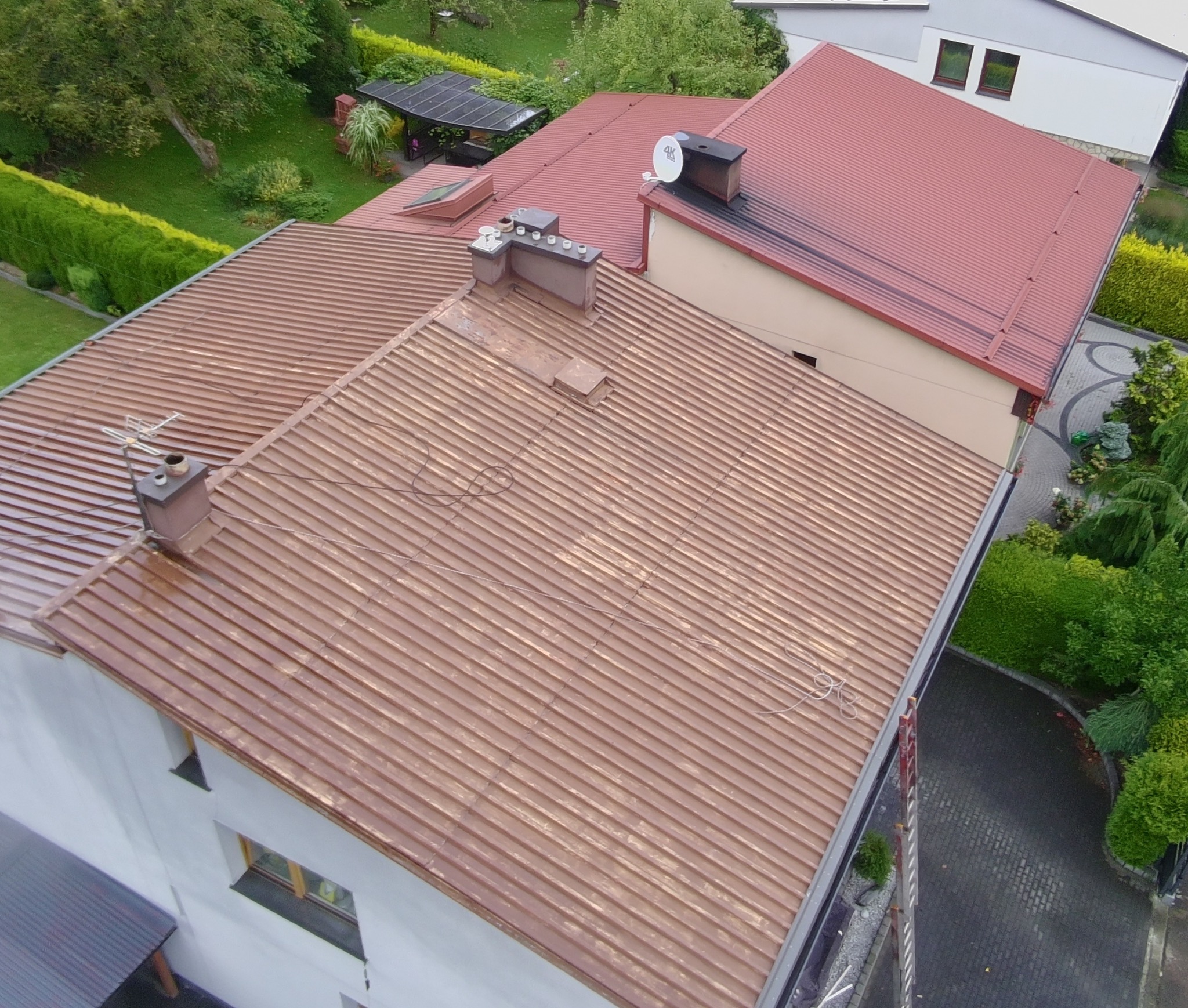 Widok z góry na dach domu z blachy trapezowej w odcieniach brązu, z widocznymi kominami i anteną satelitarną. W tle zieleń drzew i krzewów oraz fragmenty innych budynków.