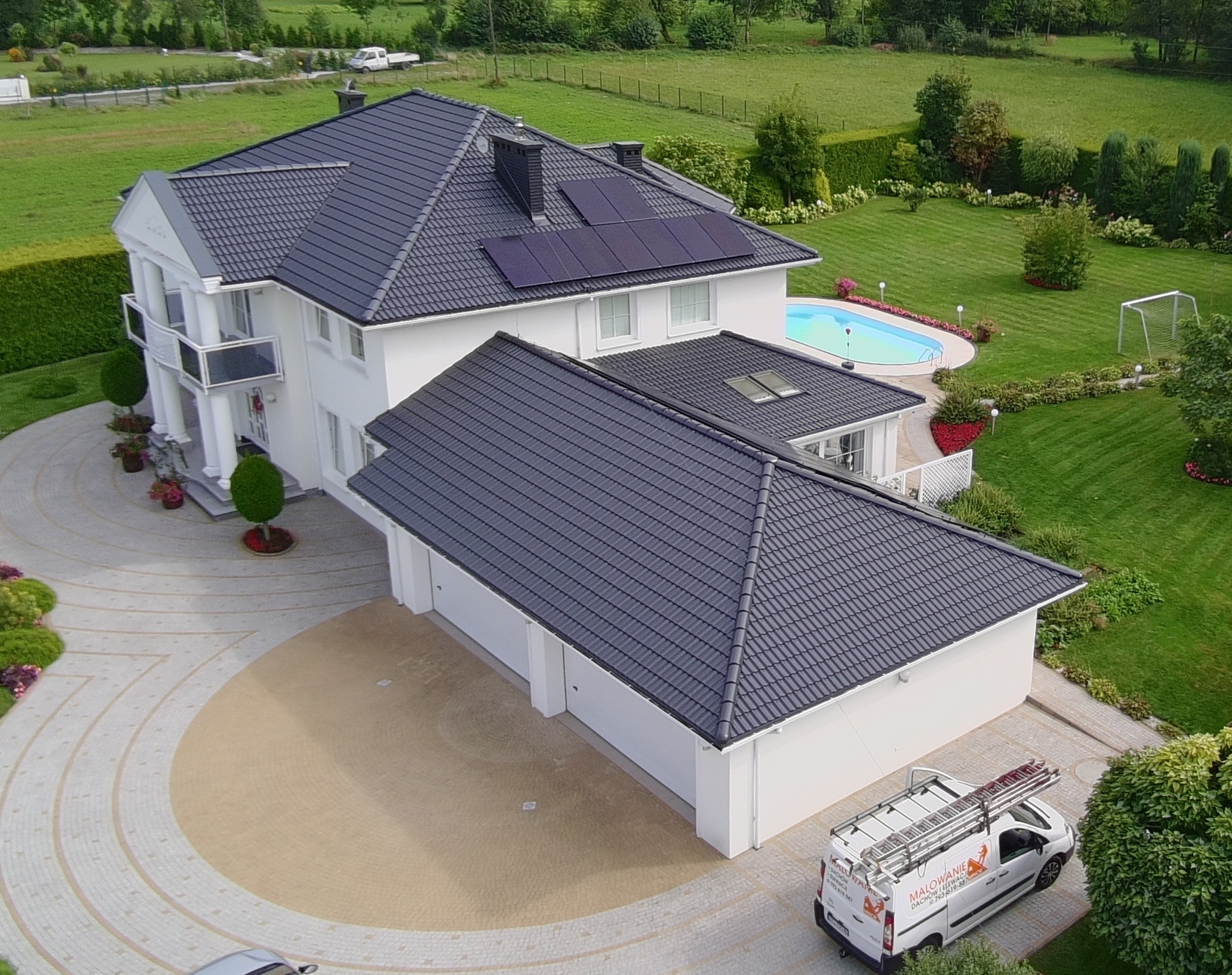 Widok z góry na dom z ciemnoszarym dachem i białymi ścianami. Na dachu panele słoneczne. Obok domu zaparkowany bus z drabiną.
