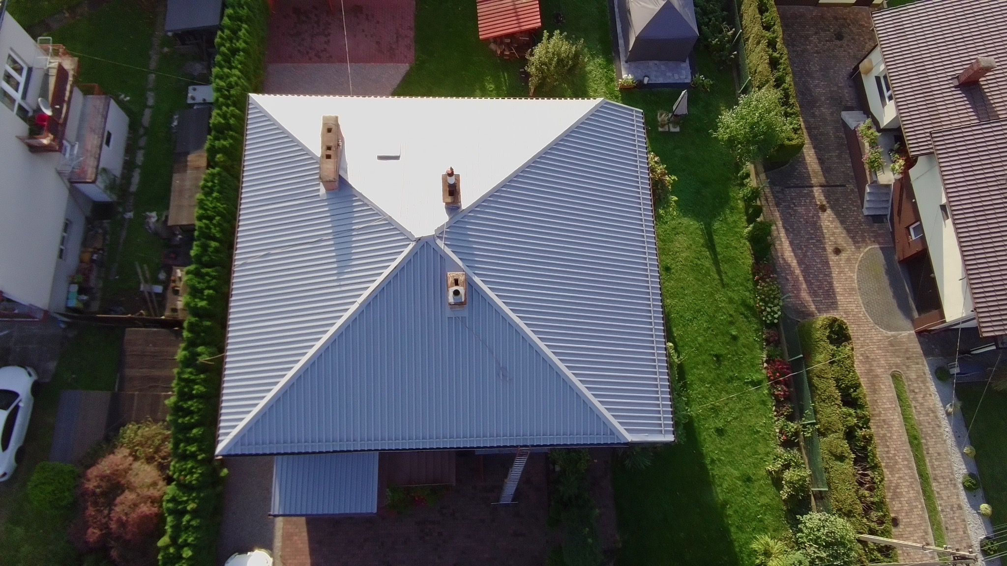Zdjęcie z drona przedstawia odnowiony, srebrny dach domu z trzema kominami, otoczony zielonym trawnikiem i drzewami. Widoczny fragment białego samochodu i sąsiednie budynki.