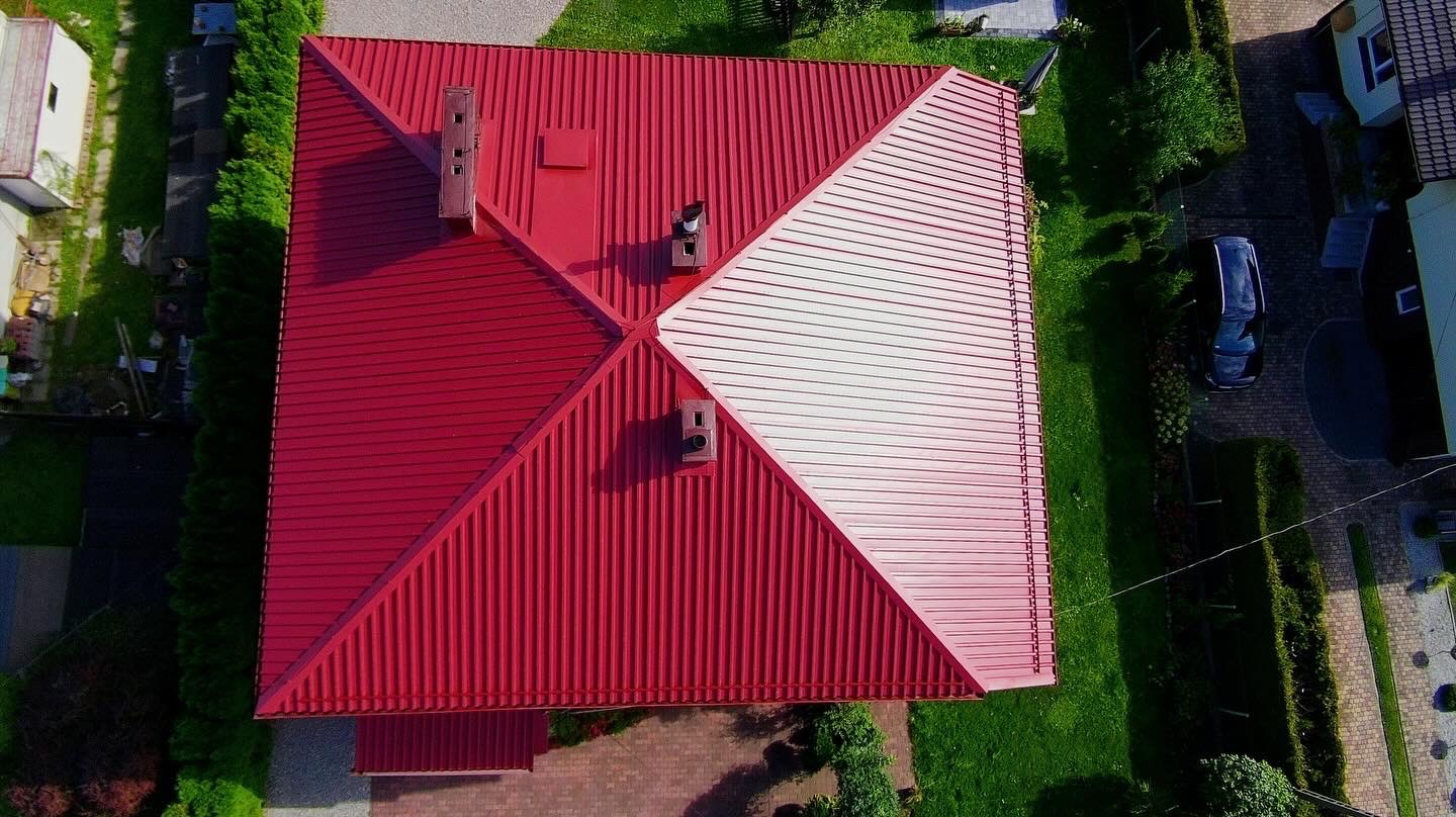 Czerwony dach z blachy trapezowej, widok z drona. Część dachu pomalowana na biało, kominy wentylacyjne. Otoczenie: trawnik, drzewa, kostka brukowa, samochód.