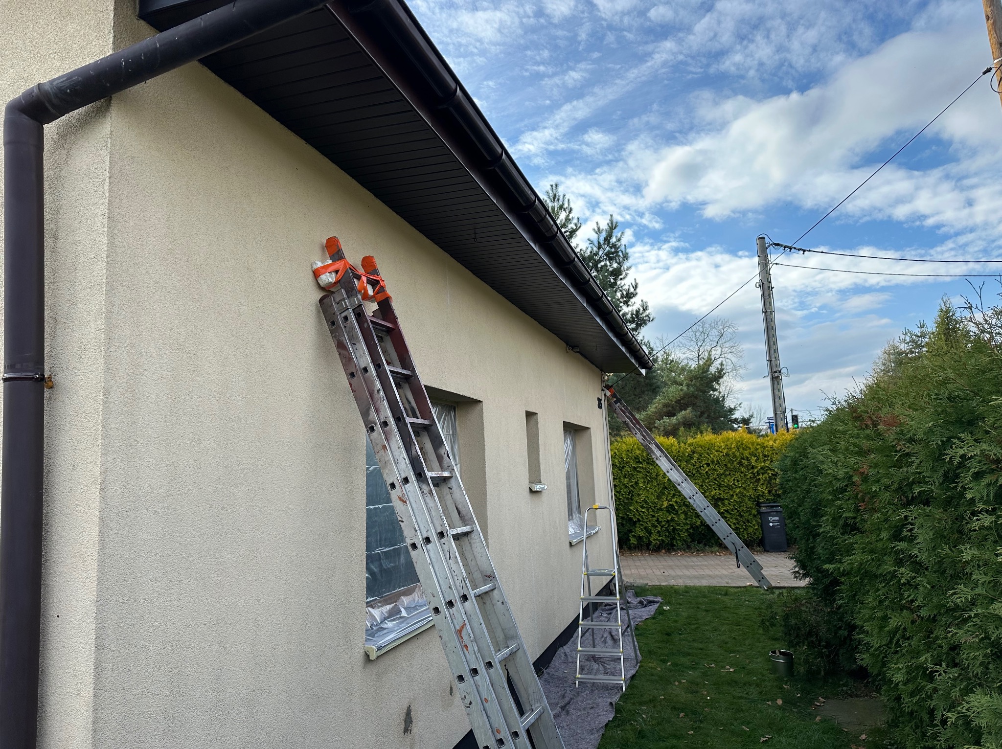 Przygotowania do malowania elewacji domu: okna zabezpieczone folią, drabiny opierają się o ścianę, widoczne rynny i fragment dachu. Słoneczny dzień.