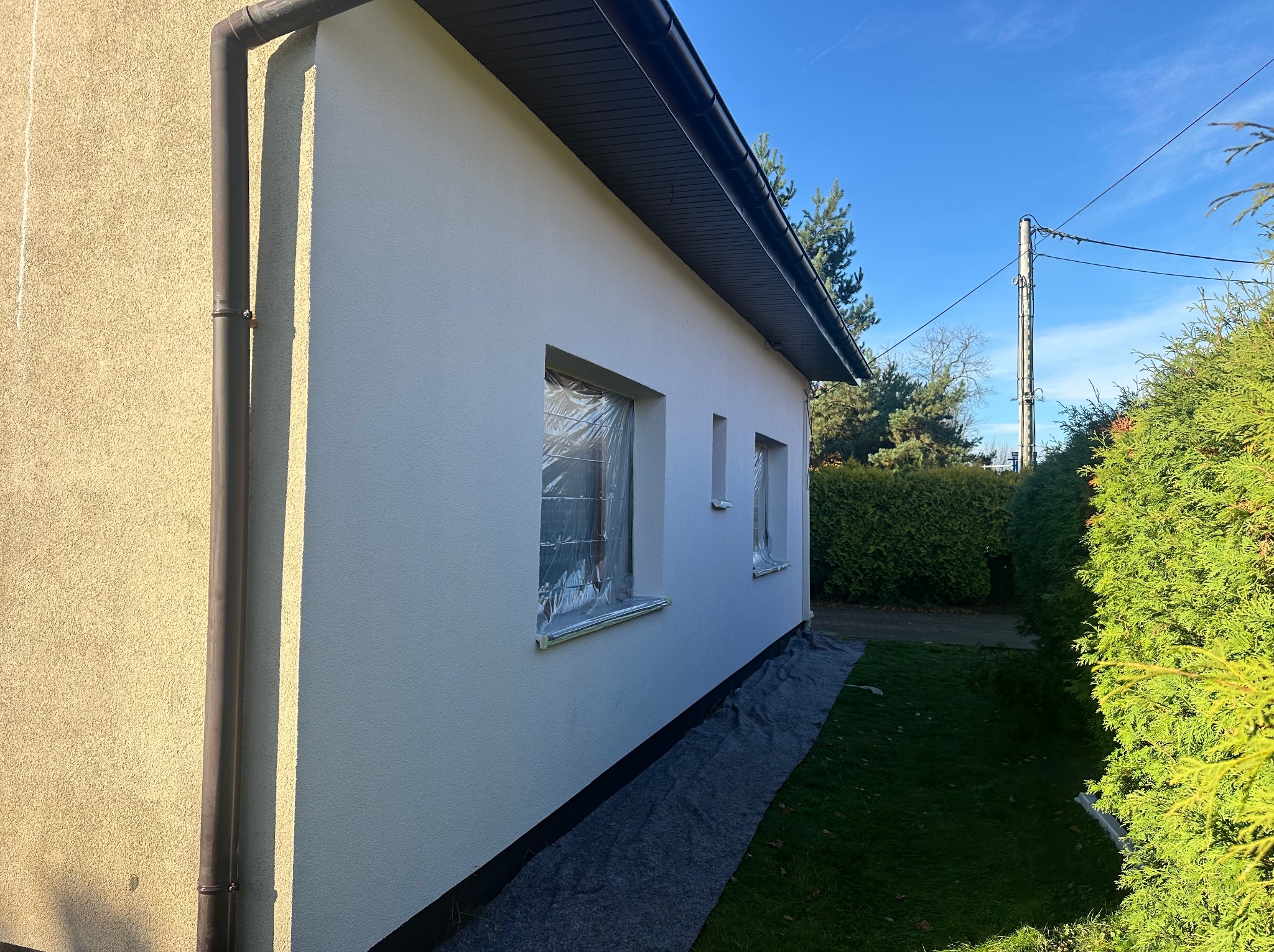 Boczny widok domu z nową, białą elewacją. Okna zabezpieczone folią malarską, widoczny fragment rynny i trawnik. Świeżo pomalowana ściana w słoneczny dzień.