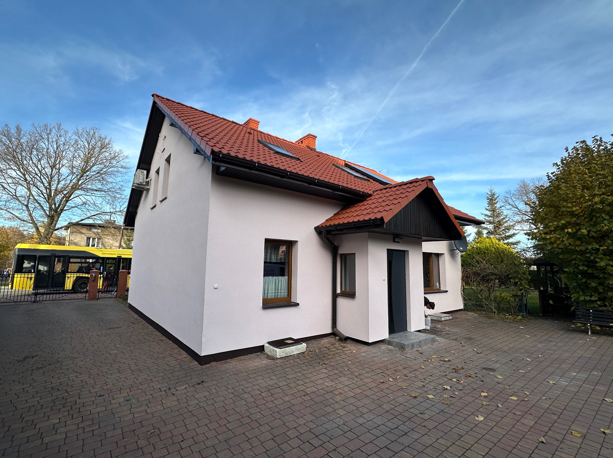 Dom jednorodzinny z czerwoną dachówką i jasną elewacją, widoczny z perspektywy chodnika z kostki brukowej. W tle drzewa i fragment żółtego autobusu.