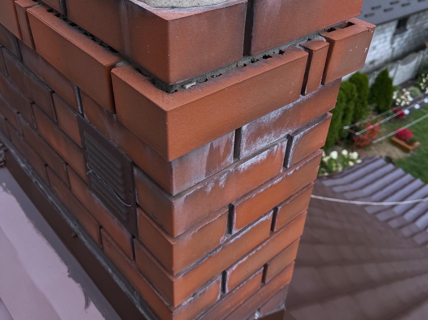 Ceglany komin z widocznymi śladami zabrudzeń i wykwitów, z metalową kratką wentylacyjną, na tle brązowego dachu i zielonego ogrodu. Ujęcie z dołu.