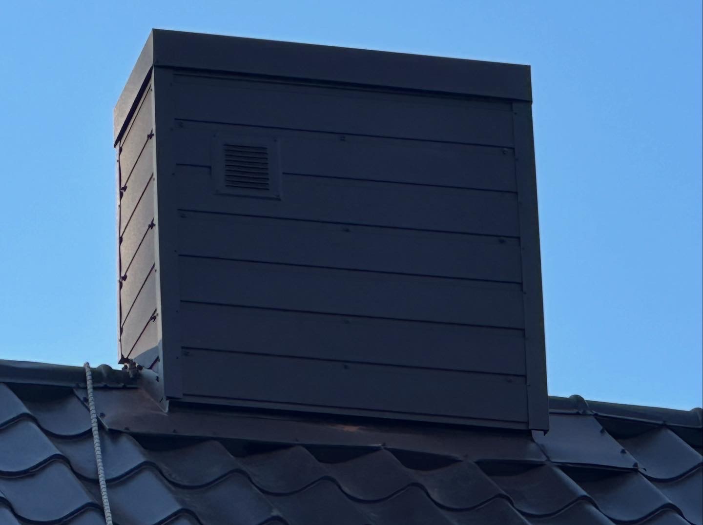 Ciemny, odnowiony komin z wentylacją na tle błękitnego nieba, widoczny z poziomu dachu pokrytego ciemną dachówką. Komin po renowacji.