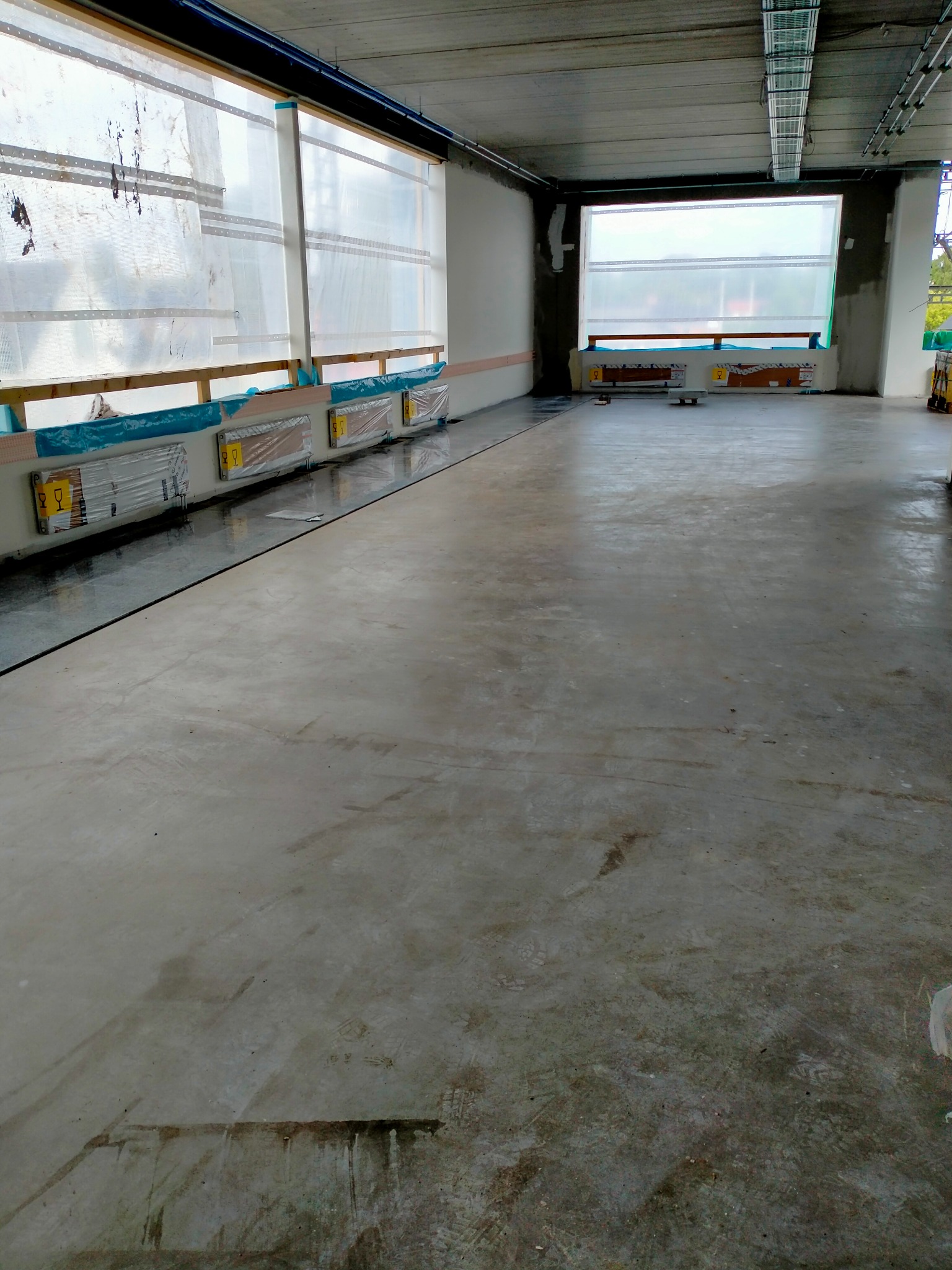 Betonowa wylewka podłoża w dużej hali, zabezpieczone okna folią, widoczne ślady po butach na powierzchni.