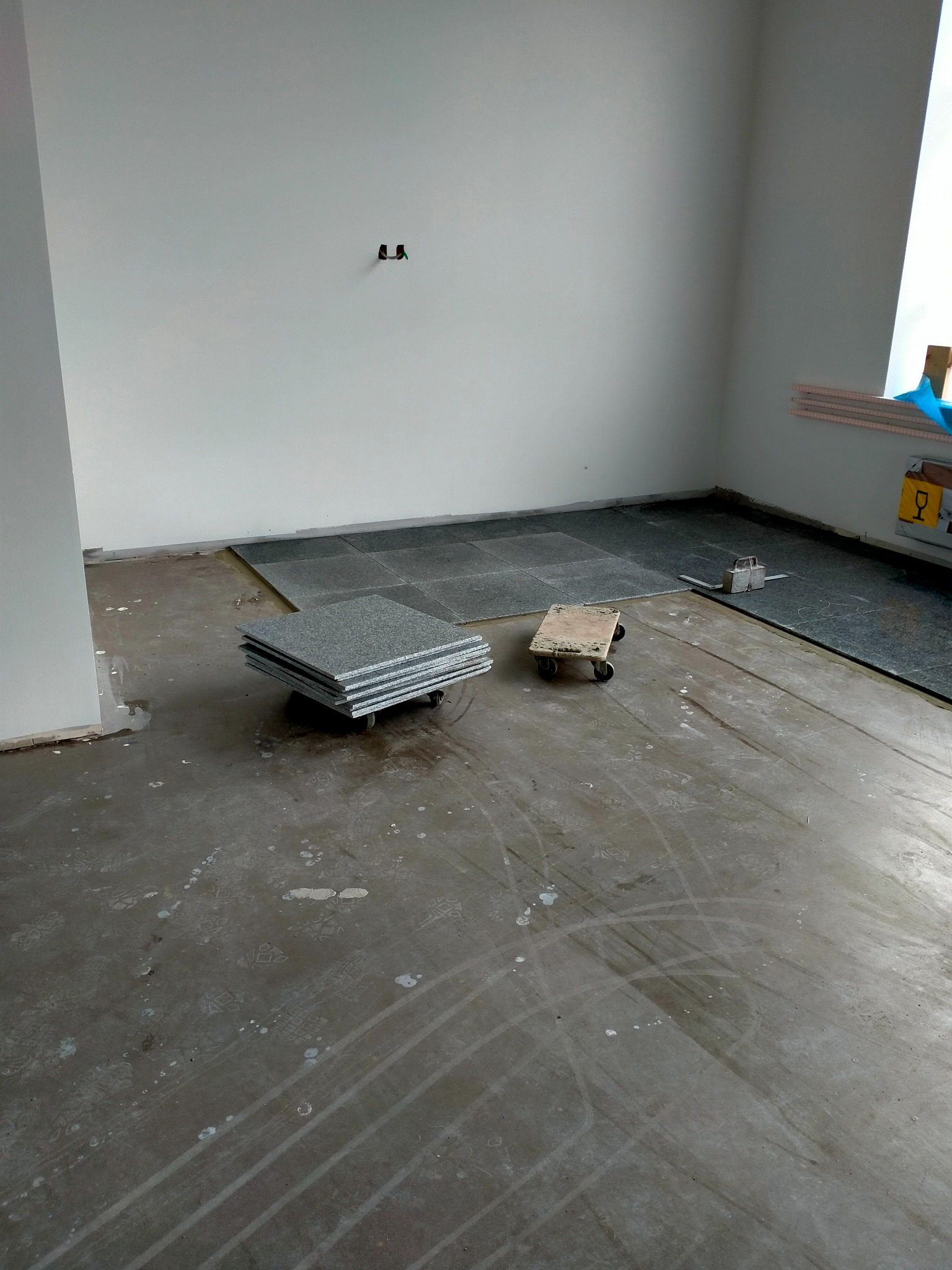 Wnętrze w trakcie układania szarych płytek posadzkowych, widoczne narzędzia i ułożone fragmenty podłogi, na podłodze ślady butów i zabrudzenia.
