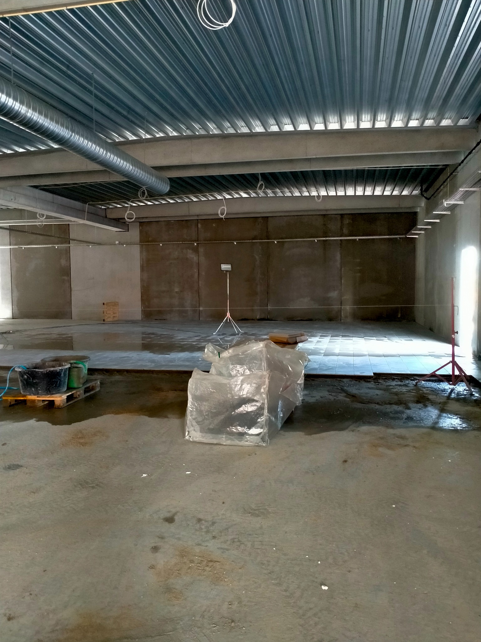 Przygotowanie podłoża pod wylewkę: mokra posadzka, wiaderka na palecie, folia ochronna i lampa budowlana w pustym pomieszczeniu z metalowym sufitem.