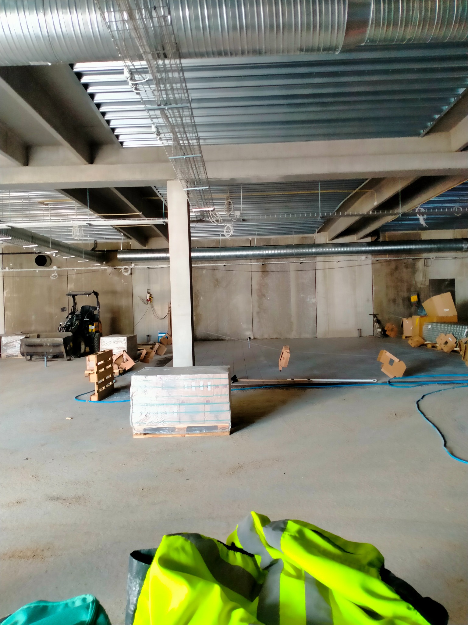 Przygotowania do wylewki posadzki przemysłowej: widoczne materiały na paletach, maszyna do wylewania, niebieski wąż i odblaskowa kamizelka na podłodze surowego wnętrza z widocznymi instalacjami...