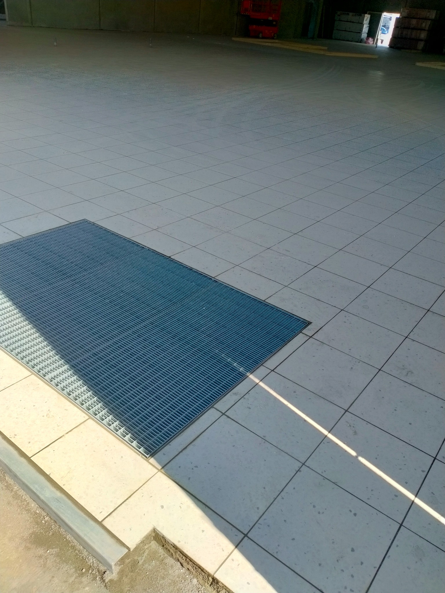 Jasnoszara, kwadratowa posadzka z kratką ściekową, oświetlona padającym promieniem słońca.