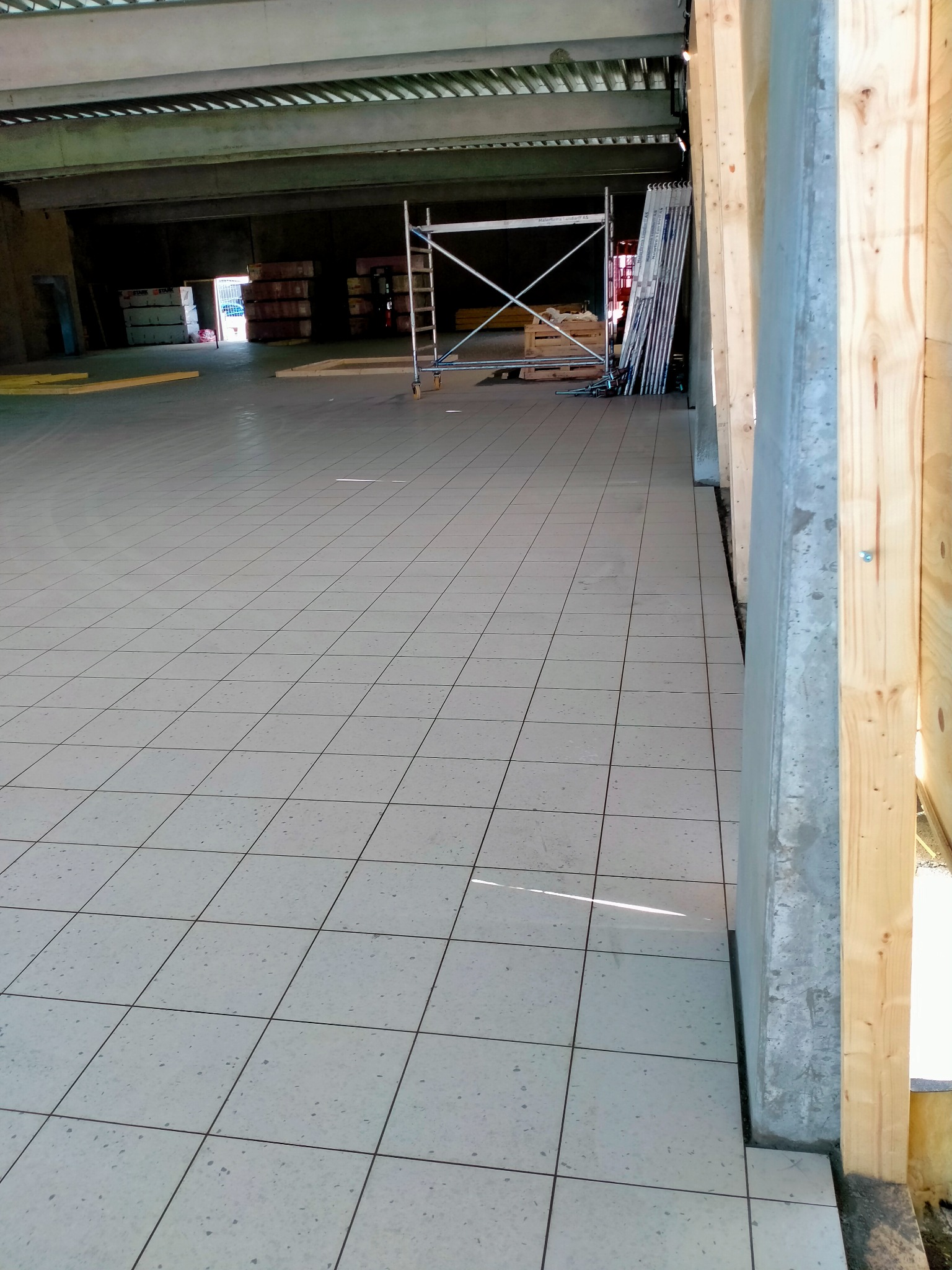 Świeżo ułożona, jasna podłoga z kwadratowych płytek w hali przemysłowej, widoczne rusztowanie i materiały budowlane w tle, drewniane elementy konstrukcyjne po prawej stronie.