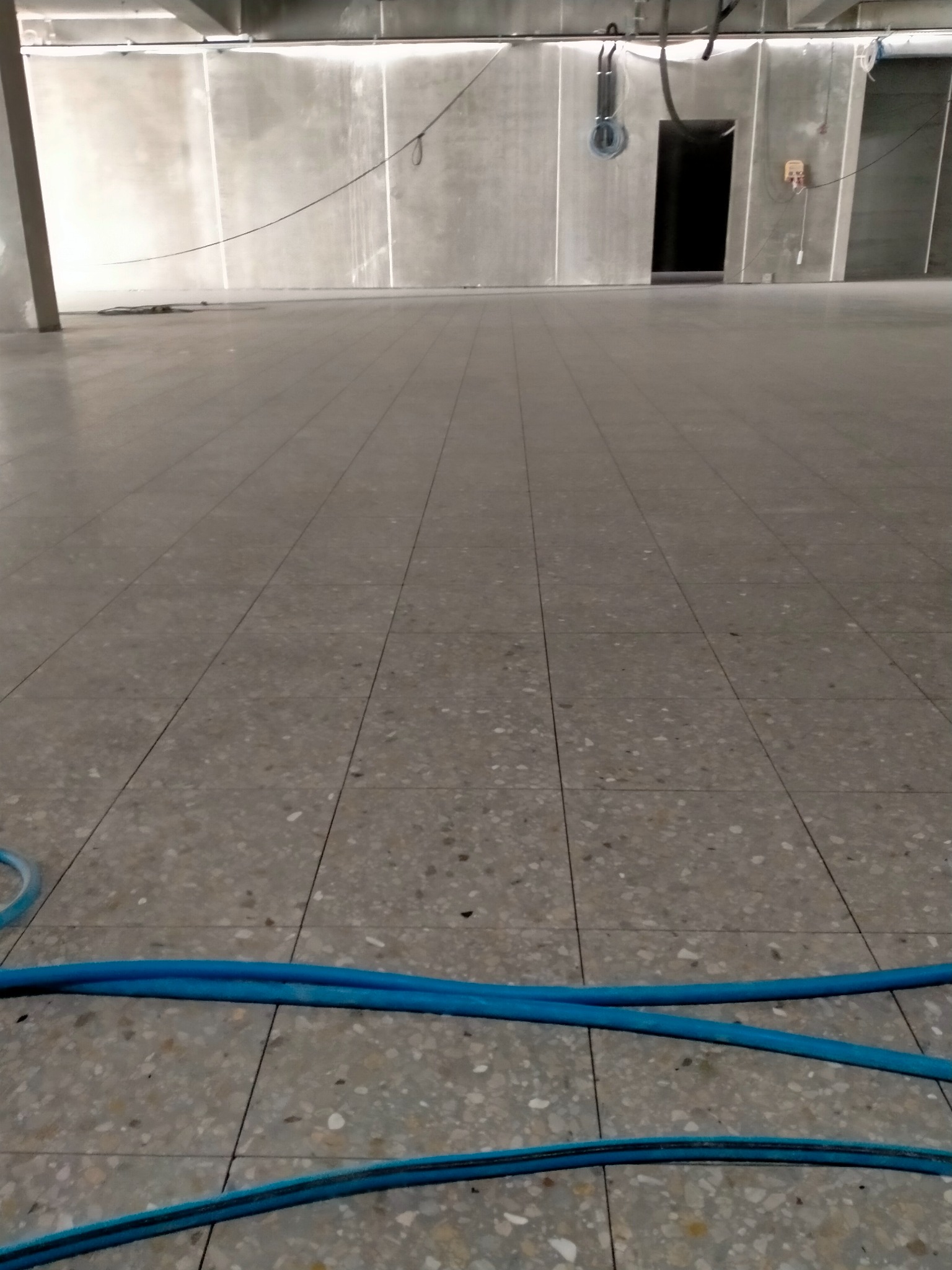 Szara, wzorzysta posadzka przemysłowa z widocznymi niebieskimi wężami na podłodze, w tle puste pomieszczenie z szarymi ścianami i otwartymi drzwiami.