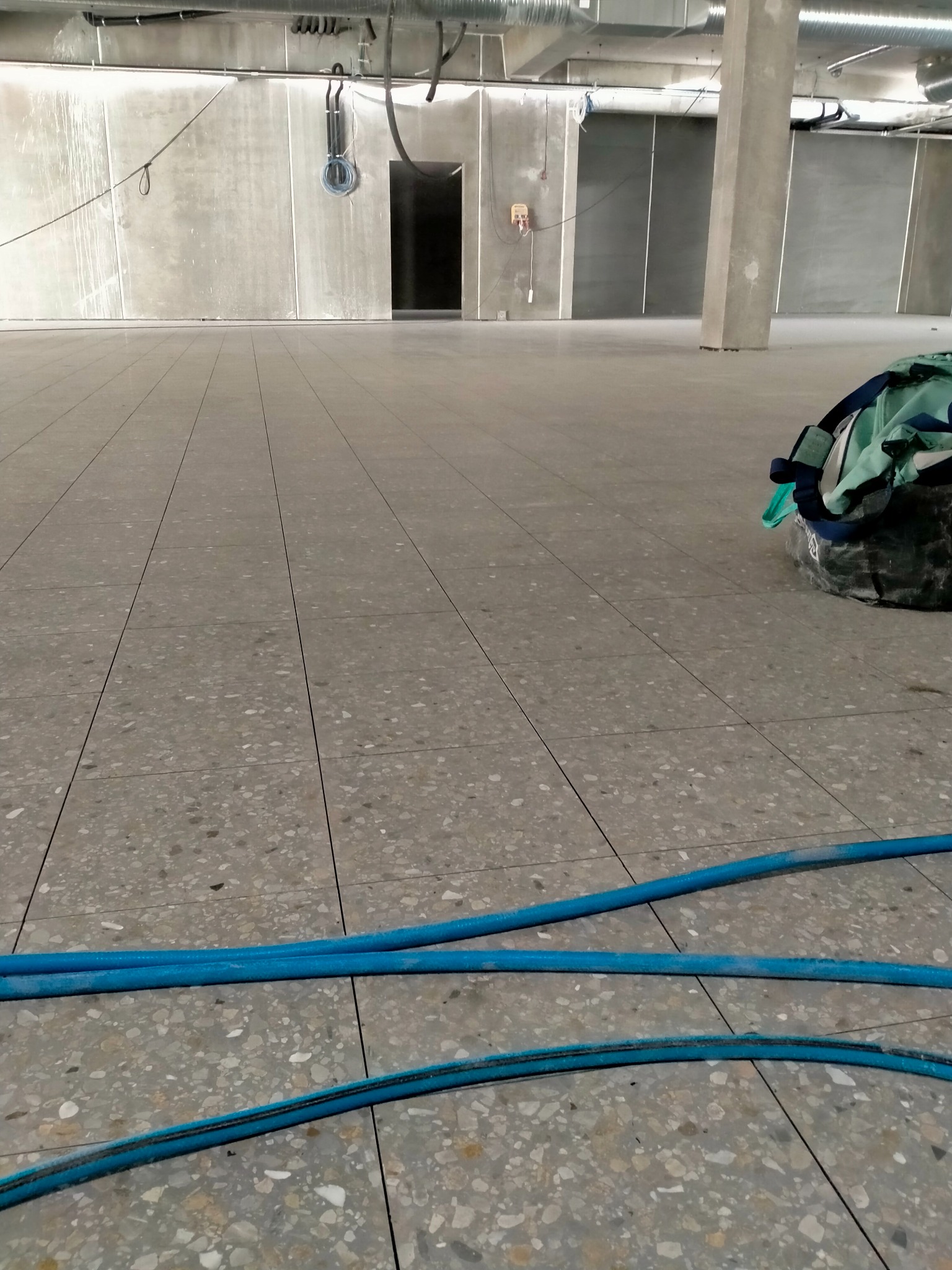 Szara, terakotowa posadzka w hali przemysłowej, widoczne niebieskie węże i torba robocza, surowe ściany i instalacje pod sufitem.