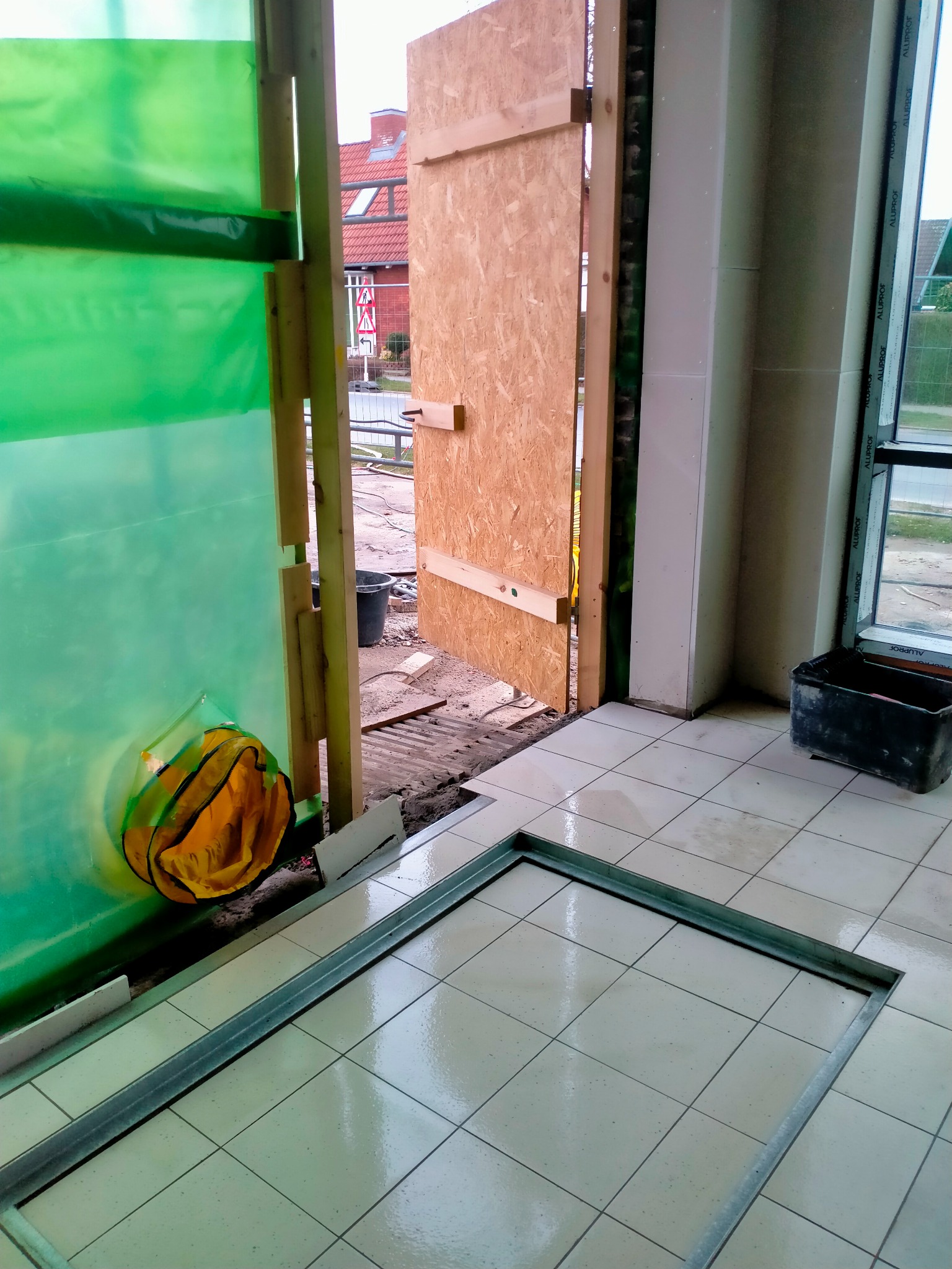 Wnętrze w Gorlicach w trakcie prac remontowych; widoczne zabezpieczenia folią, tymczasowe drzwi z płyty OSB, oraz przygotowany fragment pod wylewkę, z ramą z profili. Na podłodze białe płytki.