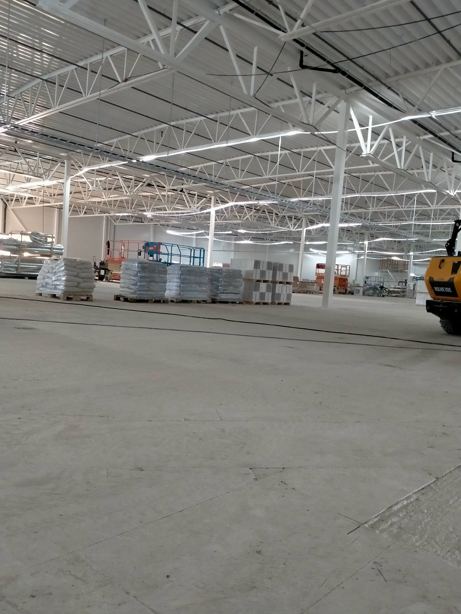 Przygotowanie do wylewki posadzki przemysłowej: palety z materiałami budowlanymi w hali z widoczną konstrukcją stalową dachu i sprzętem budowlanym w tle.