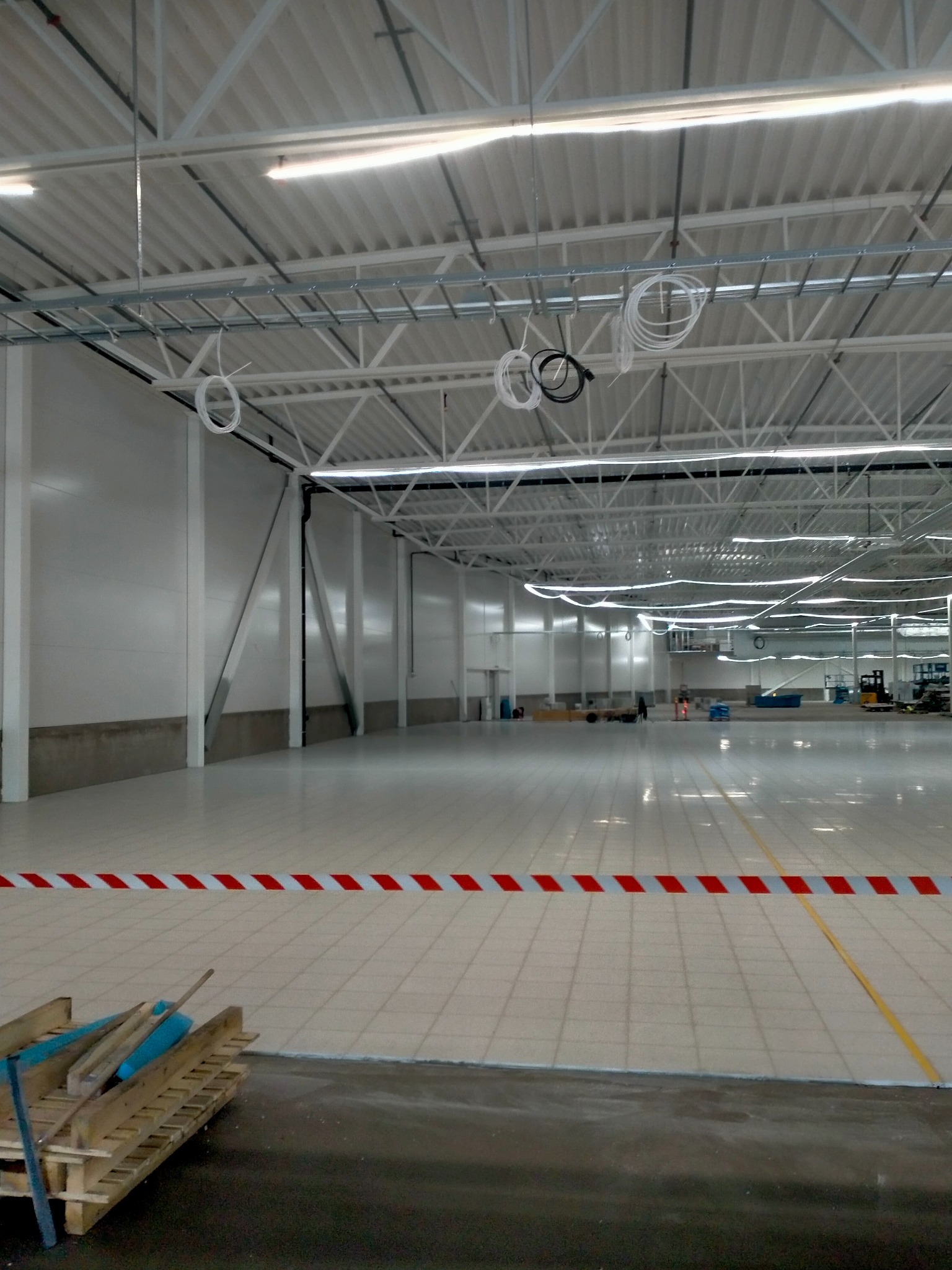 Rozległa hala przemysłowa z nową, jasną, wykafelkowaną posadzką, zabezpieczoną taśmą ostrzegawczą w biało-czerwone pasy i paletą z narzędziami w rogu; widoczne instalacje pod sufitem i maszyny...