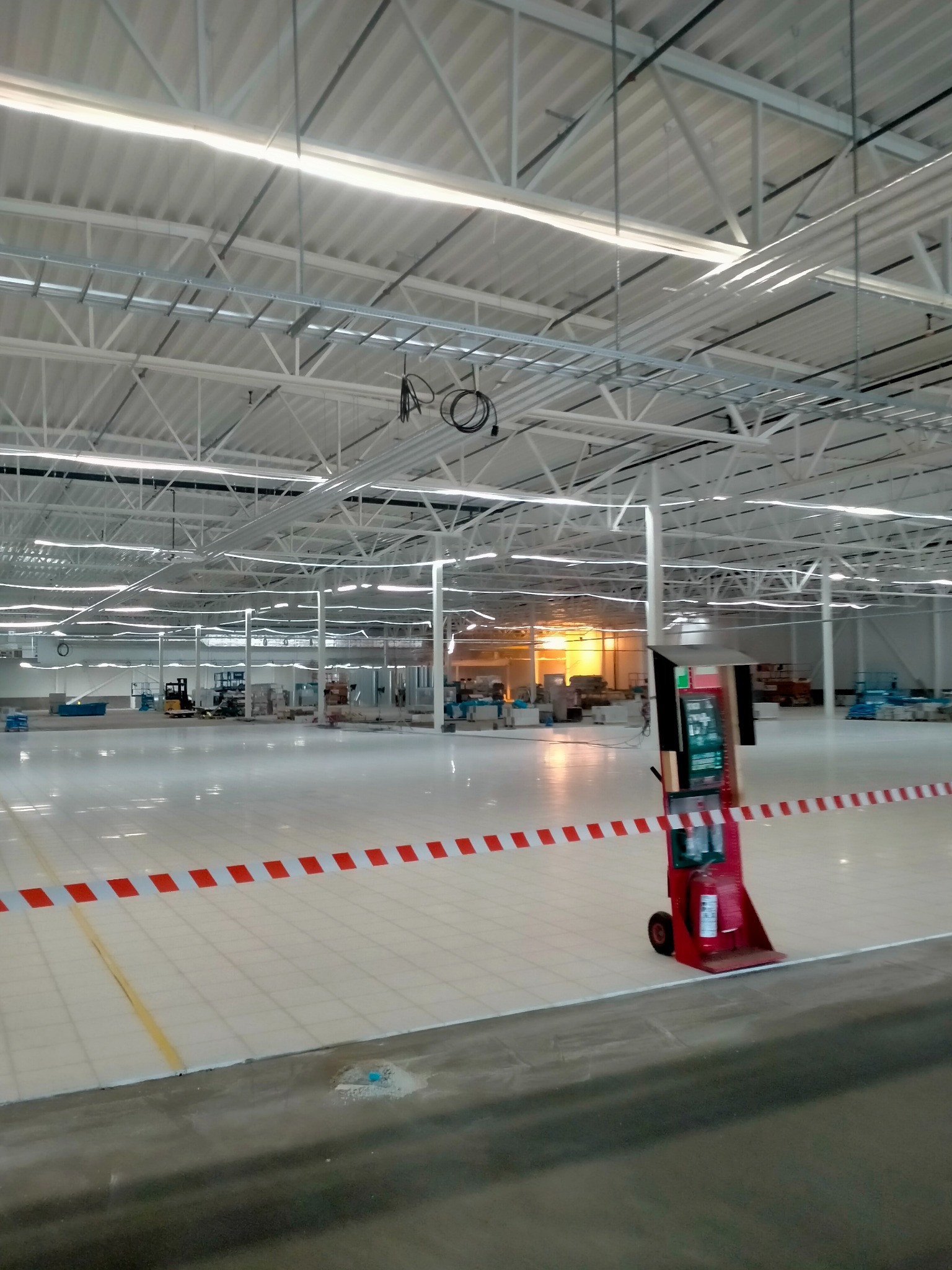 Rozległa hala przemysłowa z nową, błyszczącą posadzką w jasnym odcieniu, wyznaczona taśmą ostrzegawczą w biało-czerwone pasy, widoczny wózek z gaśnicą oraz w oddali sprzęt budowlany.