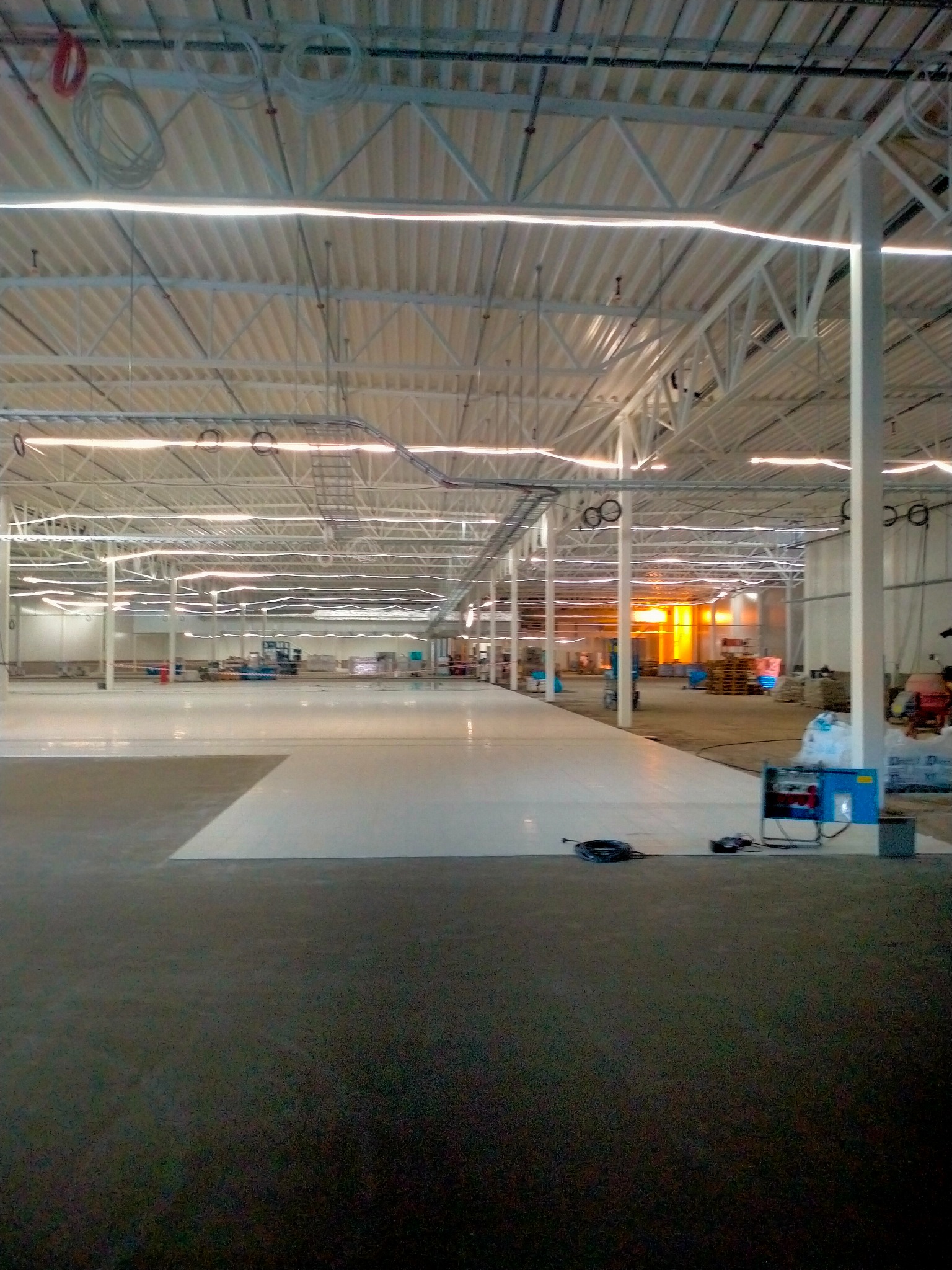 Wnętrze hali przemysłowej z częściowo wylaną, jasną posadzką, widoczne stalowe konstrukcje dachu, oświetlenie liniowe, sprzęt budowlany i instalacje elektryczne.