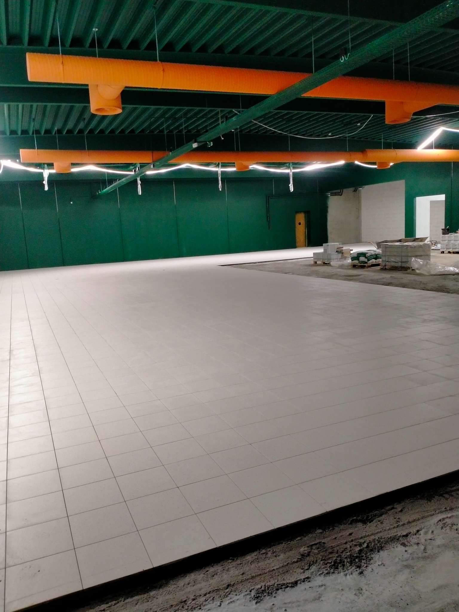Jasnoszara, kwadratowa posadzka w hali przemysłowej z zielonymi ścianami i pomarańczowymi rurami wentylacyjnymi pod sufitem. W tle widoczne materiały budowlane na paletach.