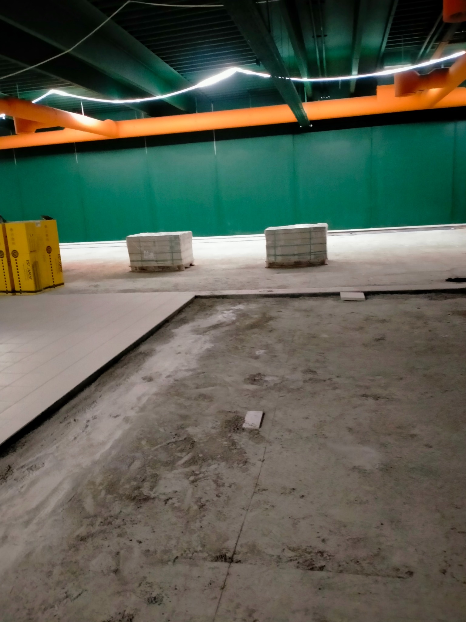 Przygotowanie podłoża pod posadzkę: widoczne ułożone płyty, palety z materiałem budowlanym i surowe, betonowe podłoże w hali z zielonymi ścianami i pomarańczowymi rurami pod sufitem.