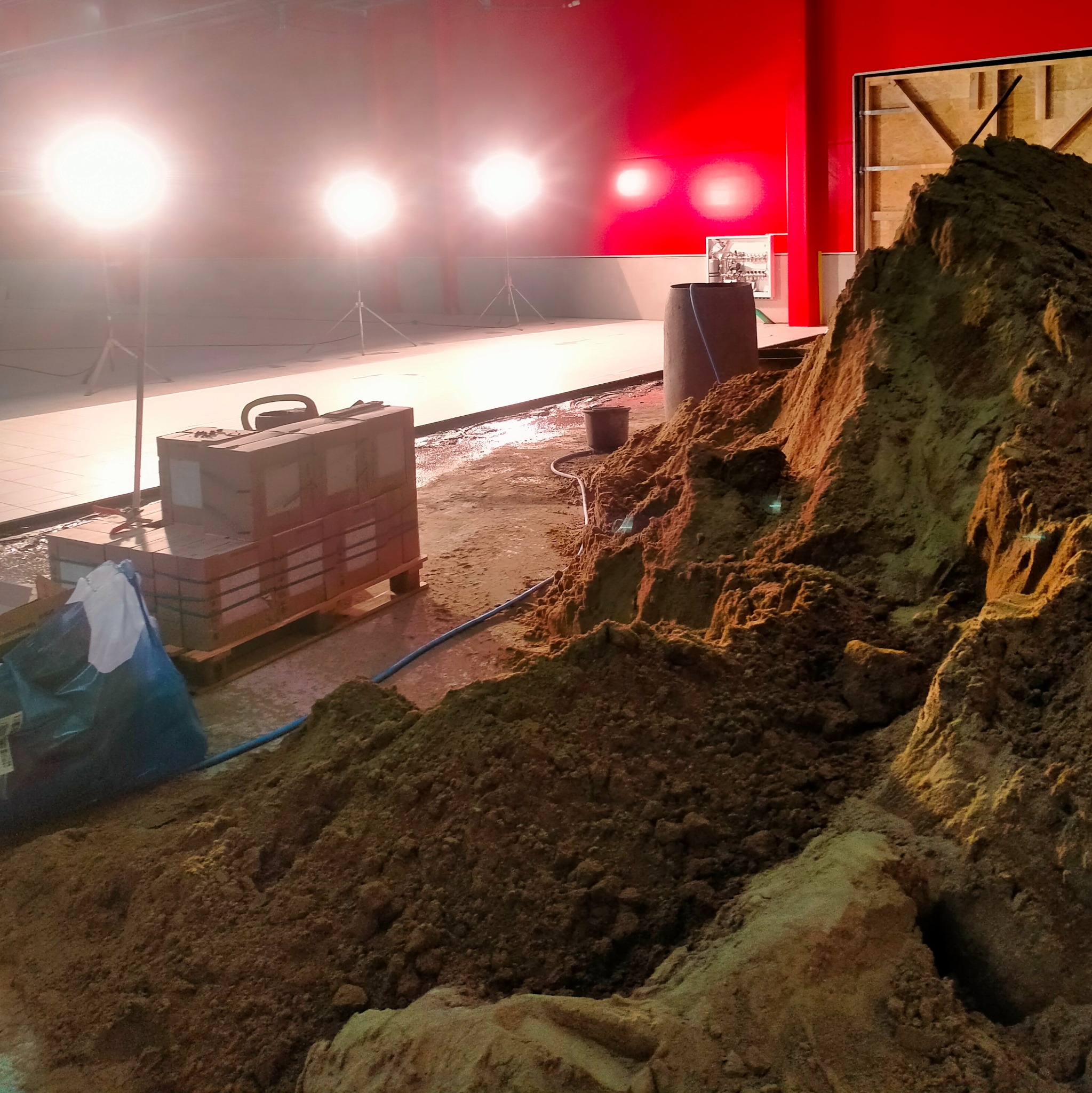 Przygotowanie podłoża pod wylewkę: Stos piasku w hali z czerwonymi ścianami, oświetlony lampami budowlanymi, obok paleta z materiałami budowlanymi i niebieski wąż.