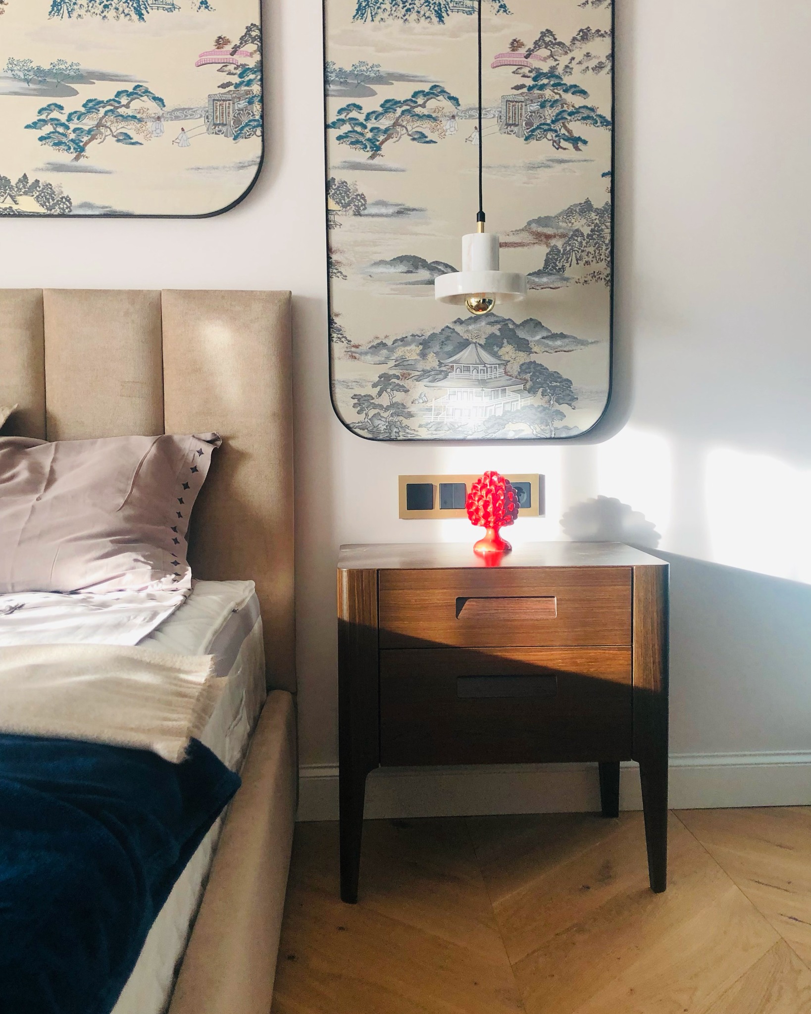 Sypialnia z łóżkiem o beżowym wezgłowiu, drewnianą szafką nocną z czerwoną dekoracją w kształcie szyszki, lampą wiszącą i panelami ściennymi z motywem azjatyckim.