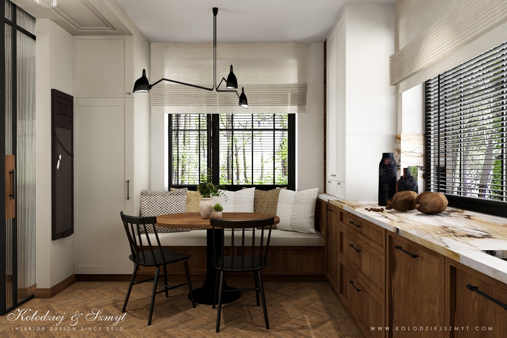 Jasna kuchnia z drewnianymi szafkami, marmurowym blatem, okrągłym stołem i wbudowaną ławką pod oknem z widokiem na zieleń, oświetlona nowoczesną lampą sufitową.