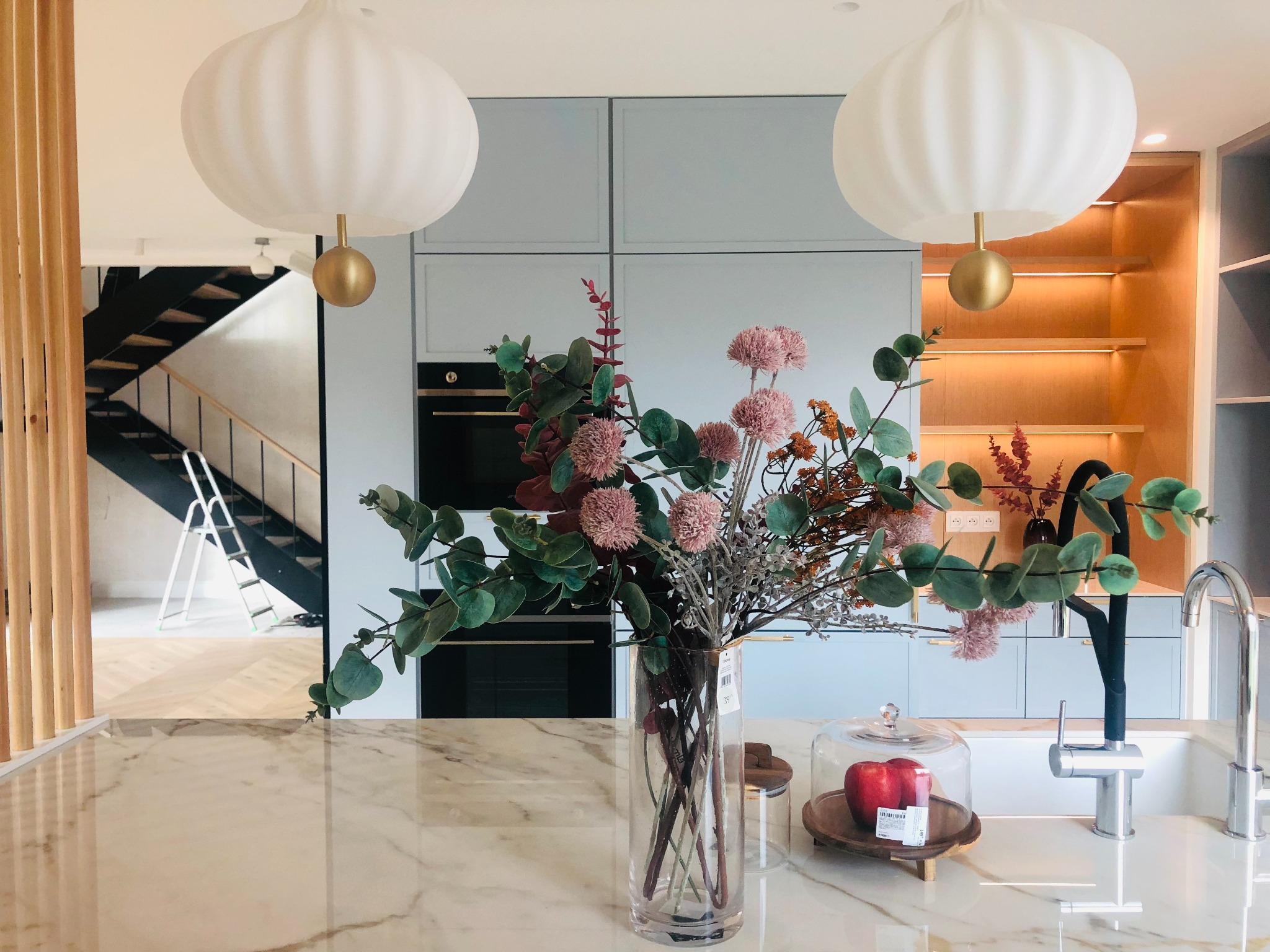 Jasna kuchnia z marmurowym blatem, na którym stoi wazon z kwiatami i jabłka pod kloszem, w tle schody z drabinką malarską.