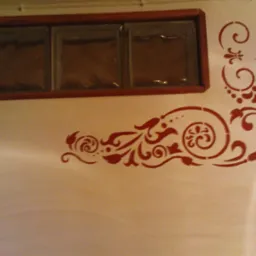 Dekoracyjny ornament w odcieniach czerwieni naniesiony na jasną ścianę, nad nim trzy szklane luksfery w drewnianej ramie.