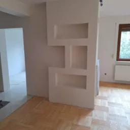 Wnętrze po remoncie z drewnianą podłogą w jodełkę i ścianą z wnękami w kolorze kremowym, widok na sąsiednie pomieszczenie z białymi płytkami podłogowymi.