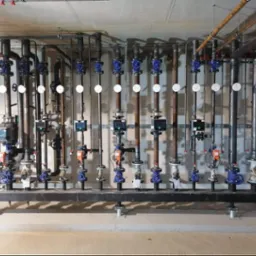 Kompleksowa instalacja grzewcza z rzędami pionowych rur miedzianych, zaworami z niebieskimi pokrętłami, manometrami i elektronicznymi regulatorami, zamontowana na czarnej belce nośnej przy...