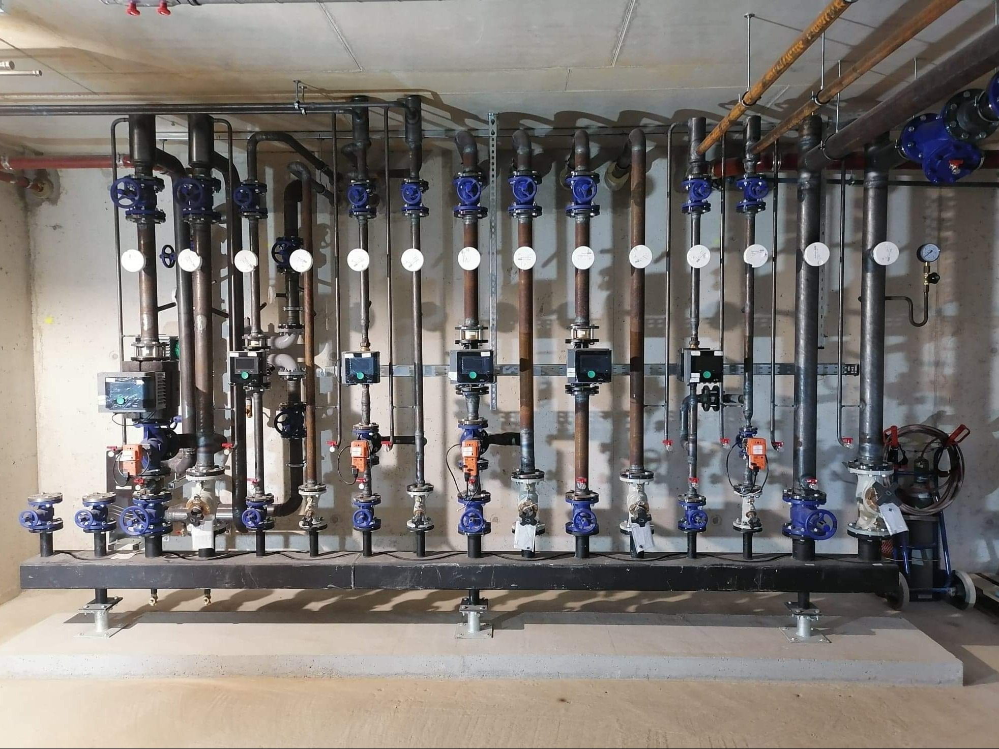 Kompleksowa instalacja grzewcza z rzędami pionowych rur miedzianych, zaworami z niebieskimi pokrętłami, manometrami i elektronicznymi regulatorami, zamontowana na czarnej belce nośnej przy...