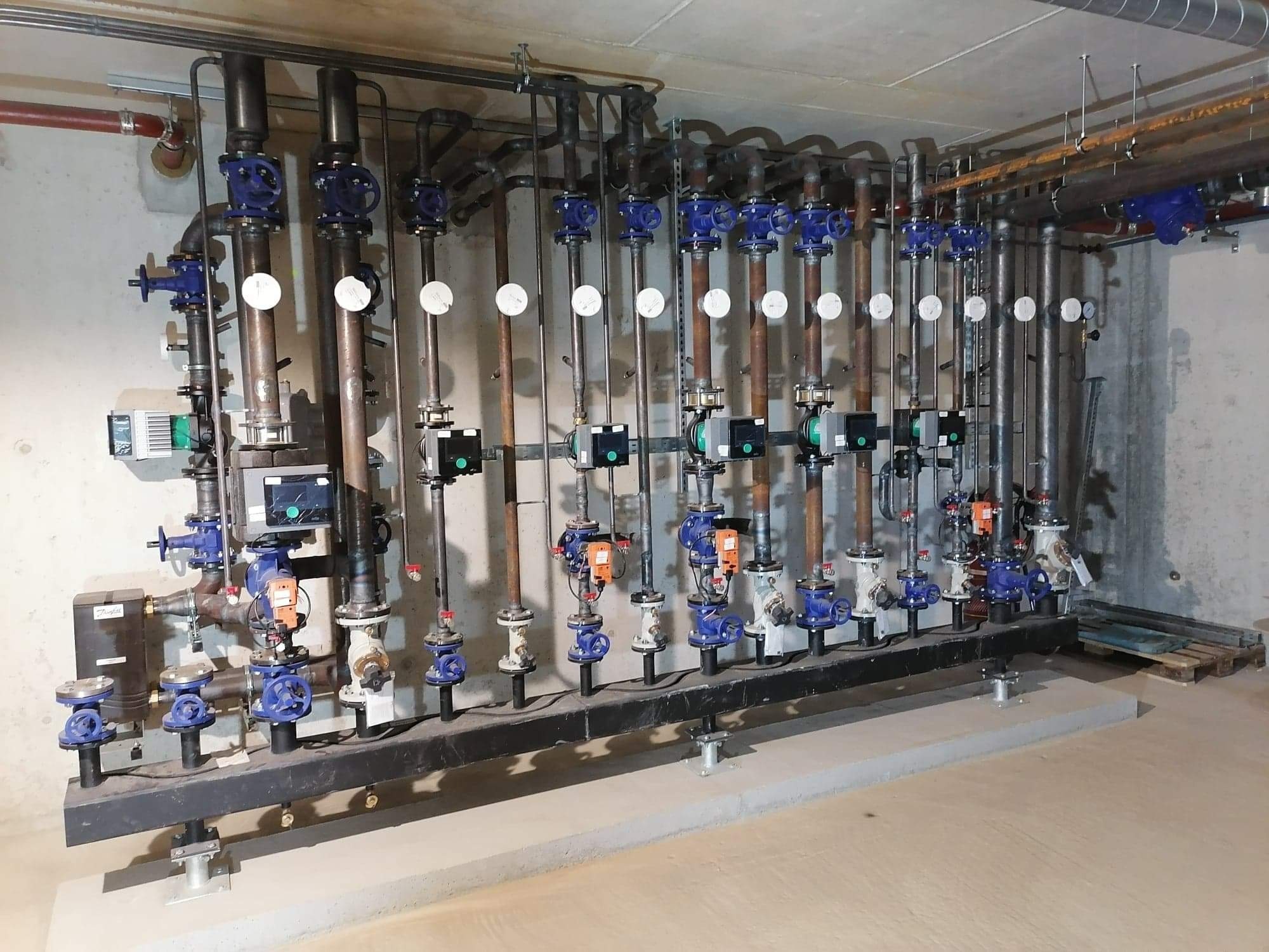 Rząd pionowych rur z zaworami i manometrami, część instalacji w pomieszczeniu technicznym z surowym betonowym wykończeniem ścian.