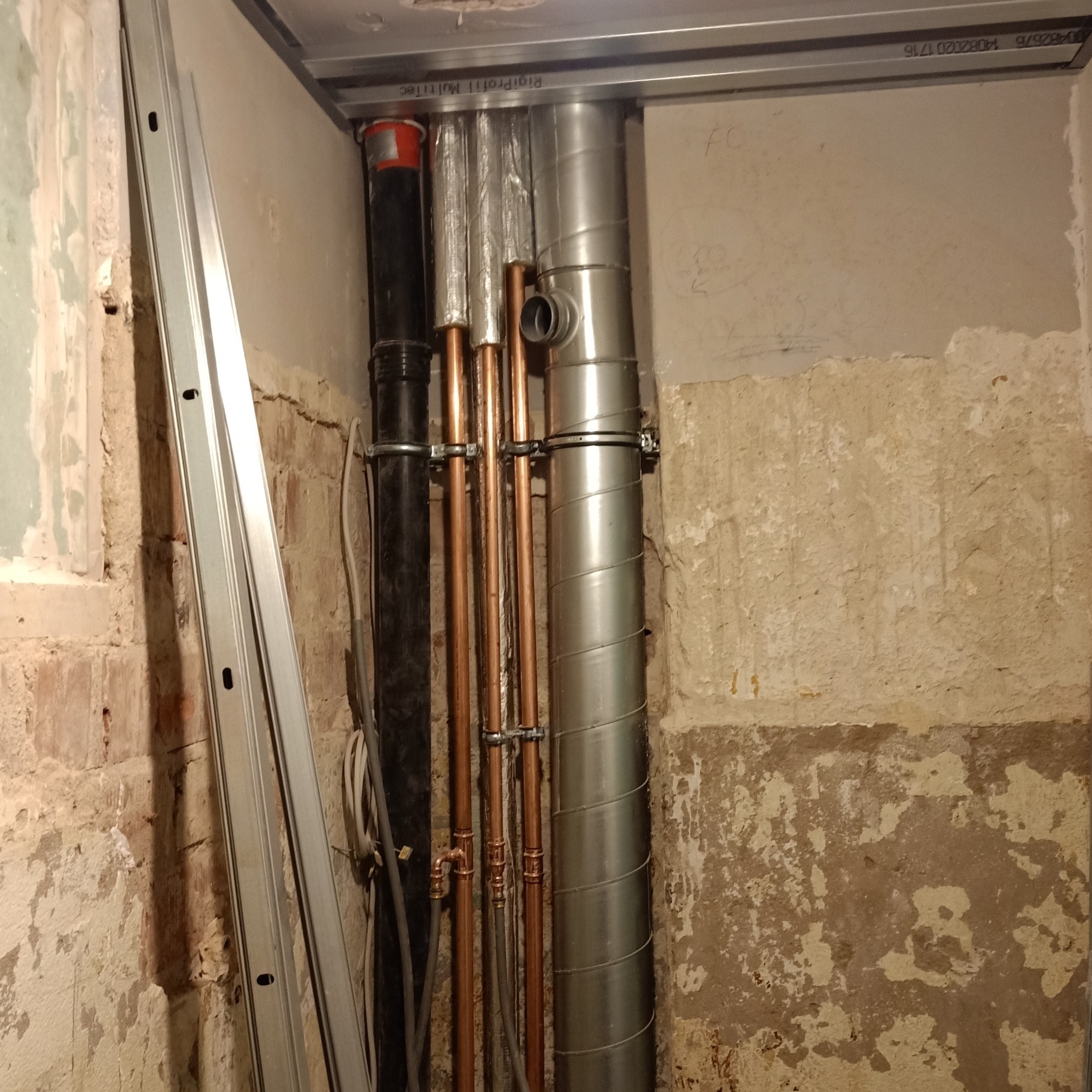 Fragment wnętrza w trakcie prac instalacyjnych: widoczne pionowe rury miedziane, czarna rura kanalizacyjna, srebrna rura wentylacyjna spiralna oraz drabina aluminiowa oparta o ścianę z widocznymi...