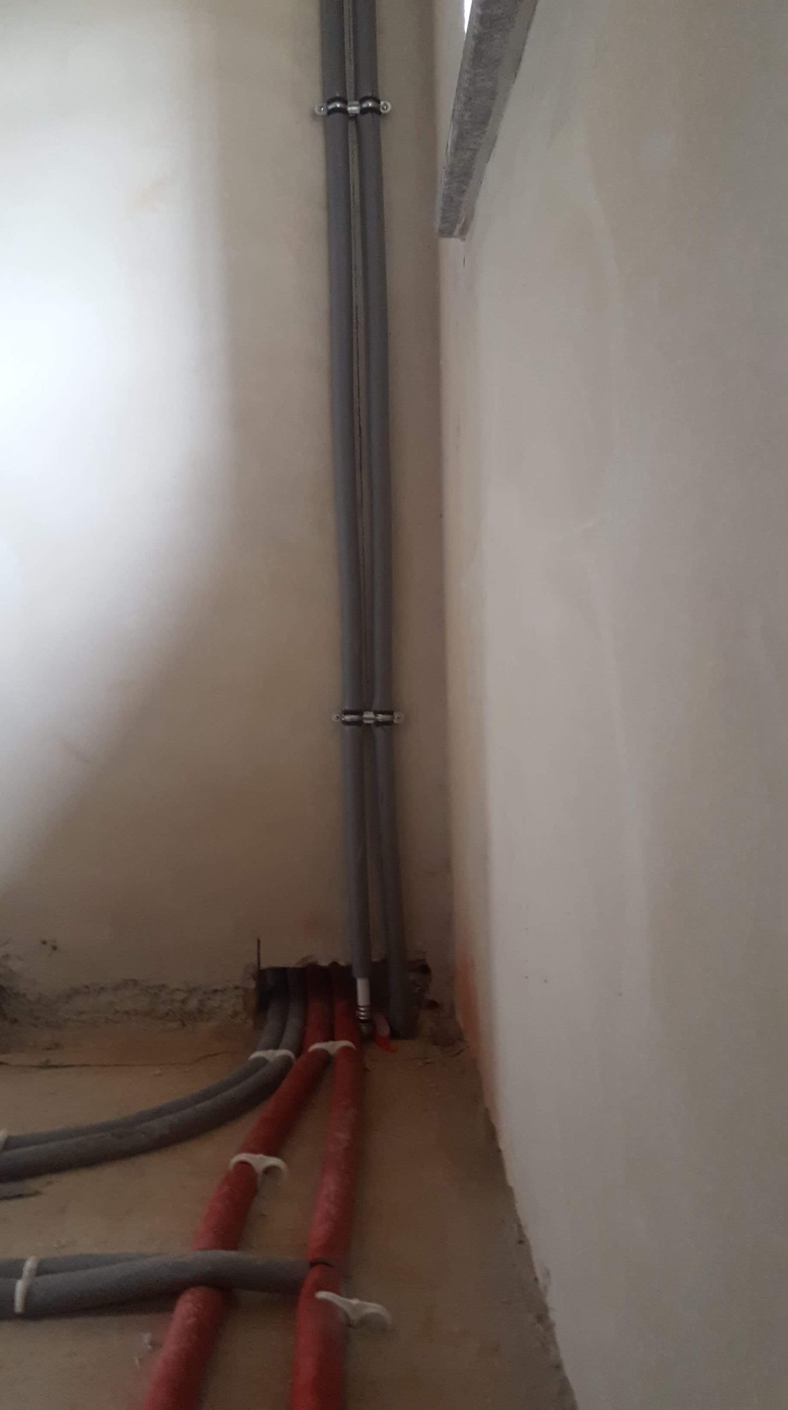 Szare i czerwone rury instalacyjne prowadzone wzdłuż ściany w surowym stanie, pionowe rury przymocowane uchwytami.