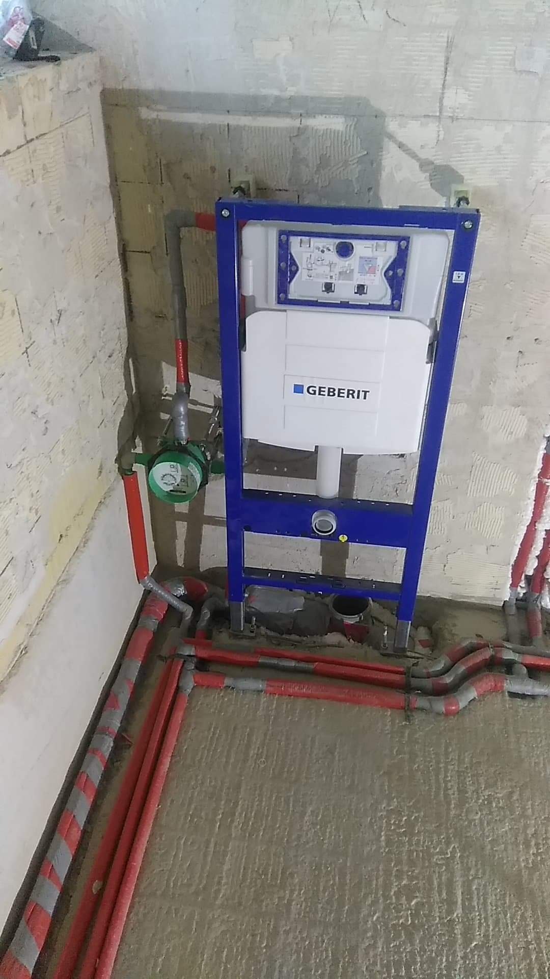 Stelaż podtynkowy Geberit zamontowany w surowym stanie, widoczne czerwone rury instalacji wodnej oraz zielony wodomierz, ściany w trakcie wykończenia.