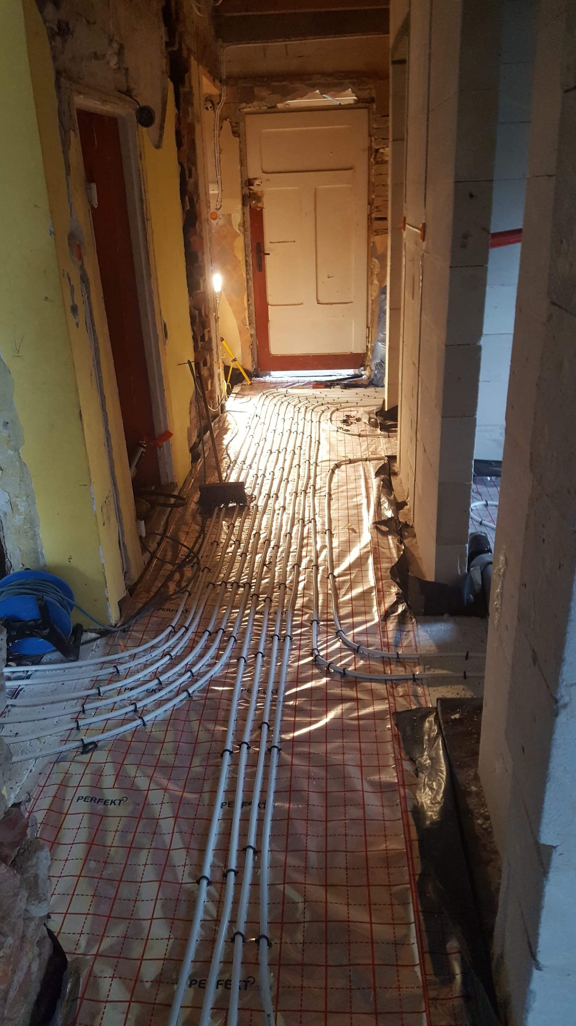 Instalacja ogrzewania podłogowego w trakcie prac: białe rurki układane na folii z czerwoną siatką w remontowanym korytarzu, widoczne ściany w stanie surowym i tymczasowe oświetlenie.