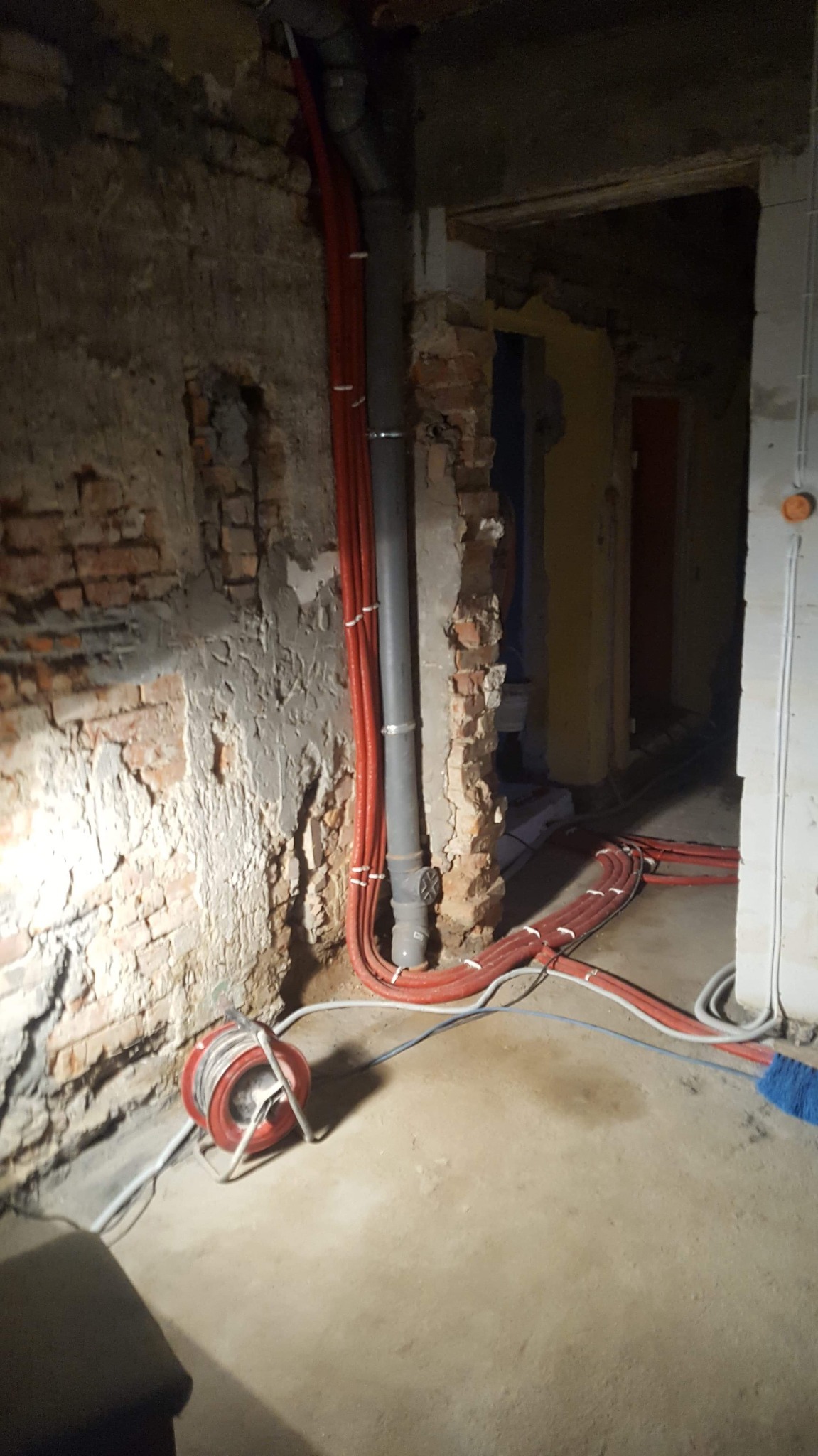 Wnętrze w trakcie remontu z widocznymi nowymi instalacjami: pion kanalizacyjny i wiązka czerwonych rur w karbowanej osłonie, zwinięty przewód na bębnie przy ścianie z odsłoniętą cegłą.
