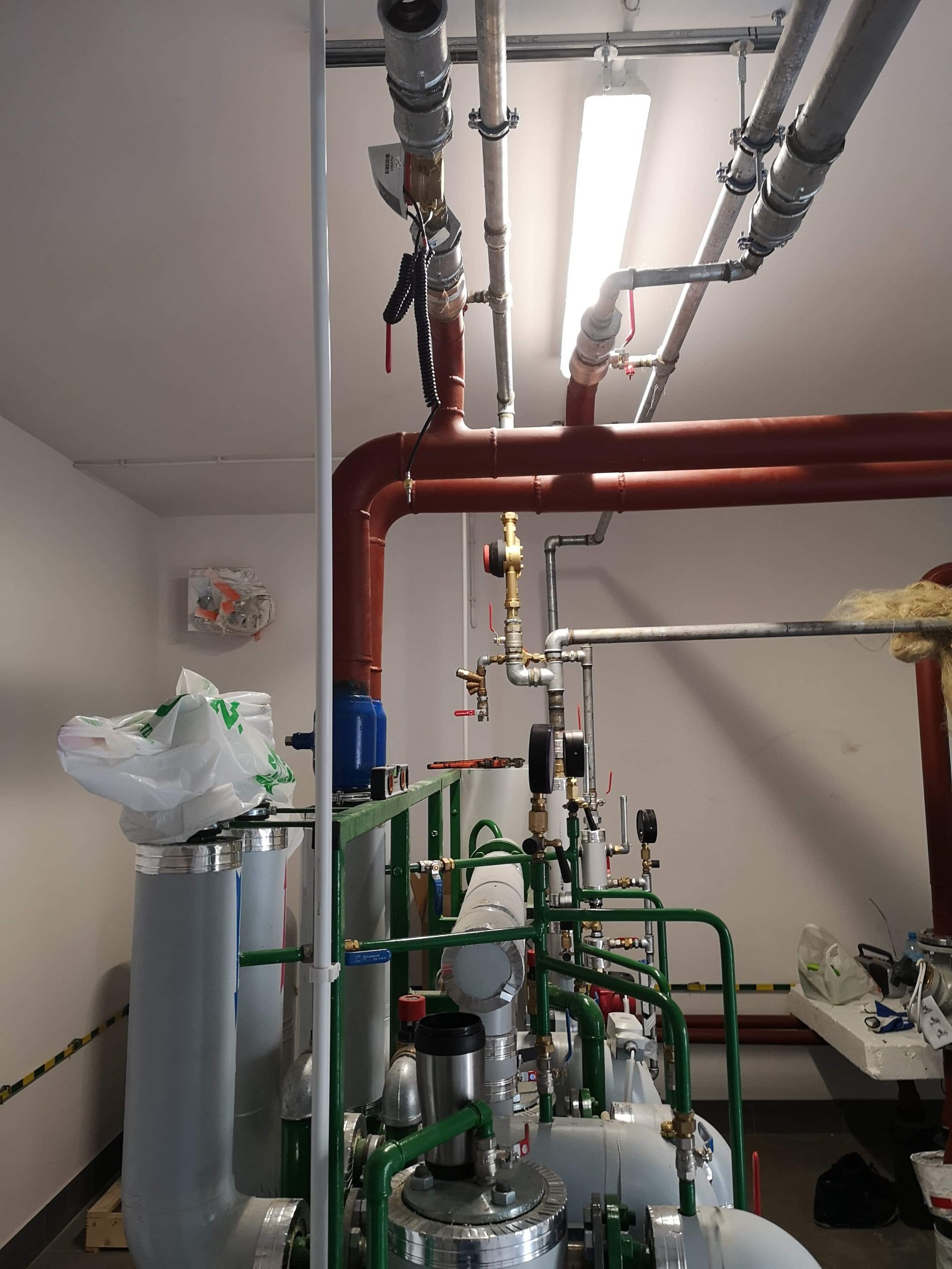Rozbudowana instalacja grzewcza z rurami miedzianymi, stalowymi i elementami armatury, widoczne filtry, manometry i zawory, całość w pomieszczeniu technicznym.