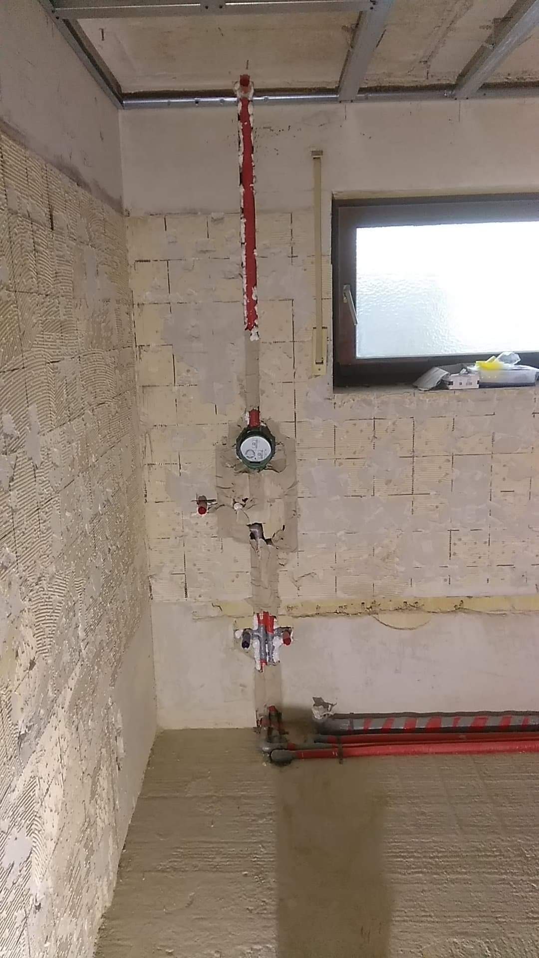 Wnętrze łazienki w trakcie remontu, widoczne czerwone rury instalacji wodnej i kanalizacyjnej, zamontowane na ścianie pokrytej klejem do płytek, oraz wylewka betonowa na podłodze, sufit z widoczną...